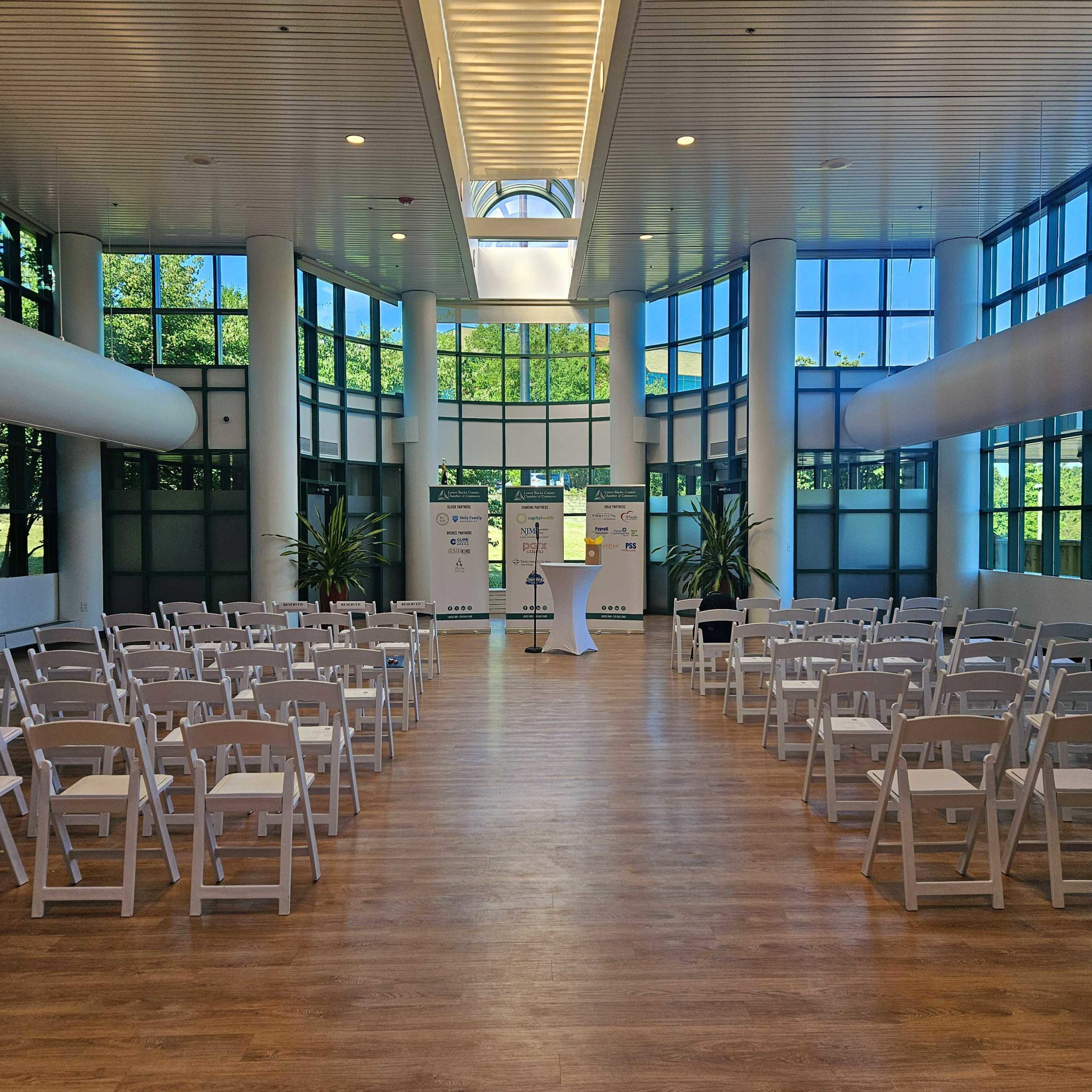 A large, bright indoor space with rows of white folding chairs facing a podium, surrounded by large windows and greenery.