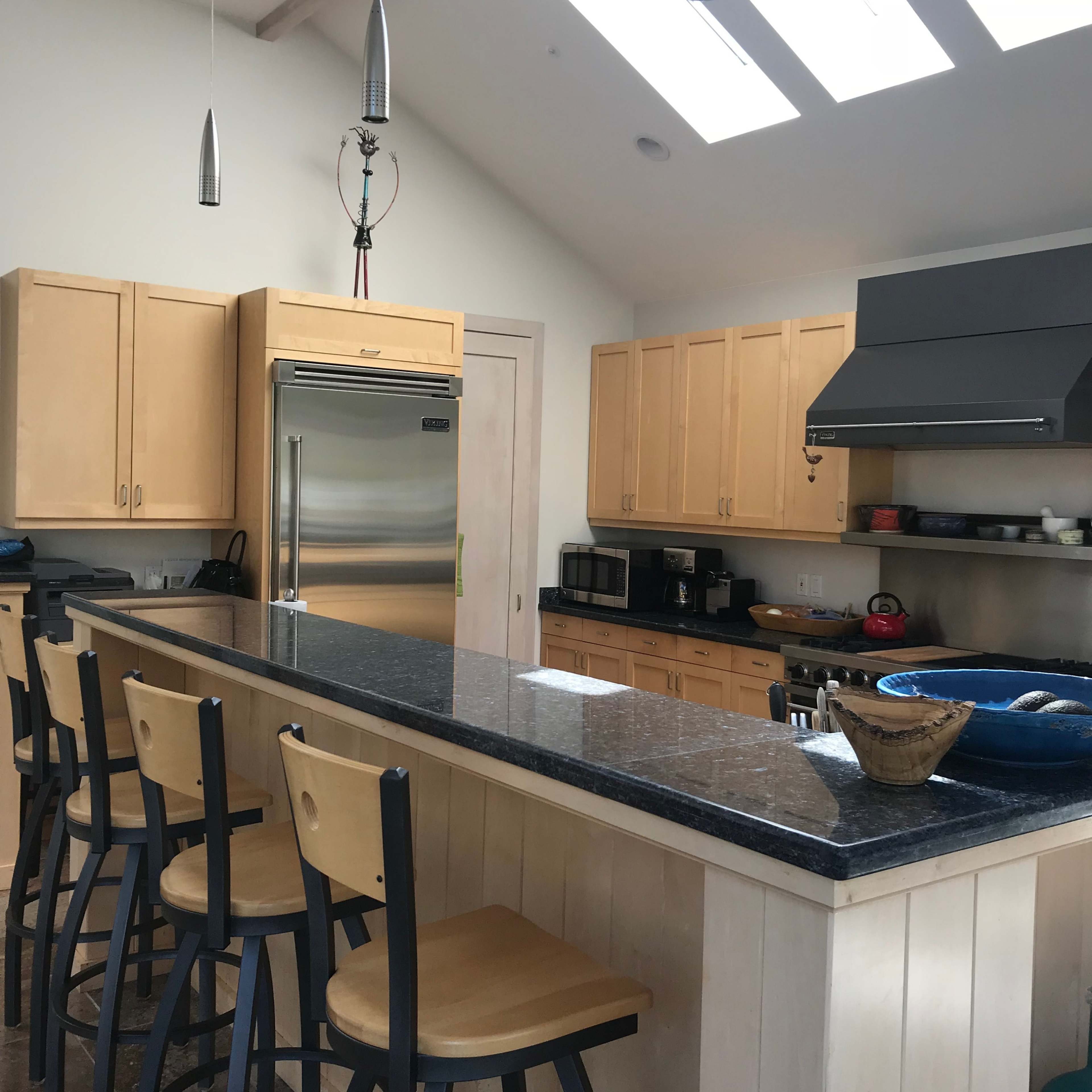 A modern kitchen features light wood cabinetry, a central island with barstools, and skylights above.