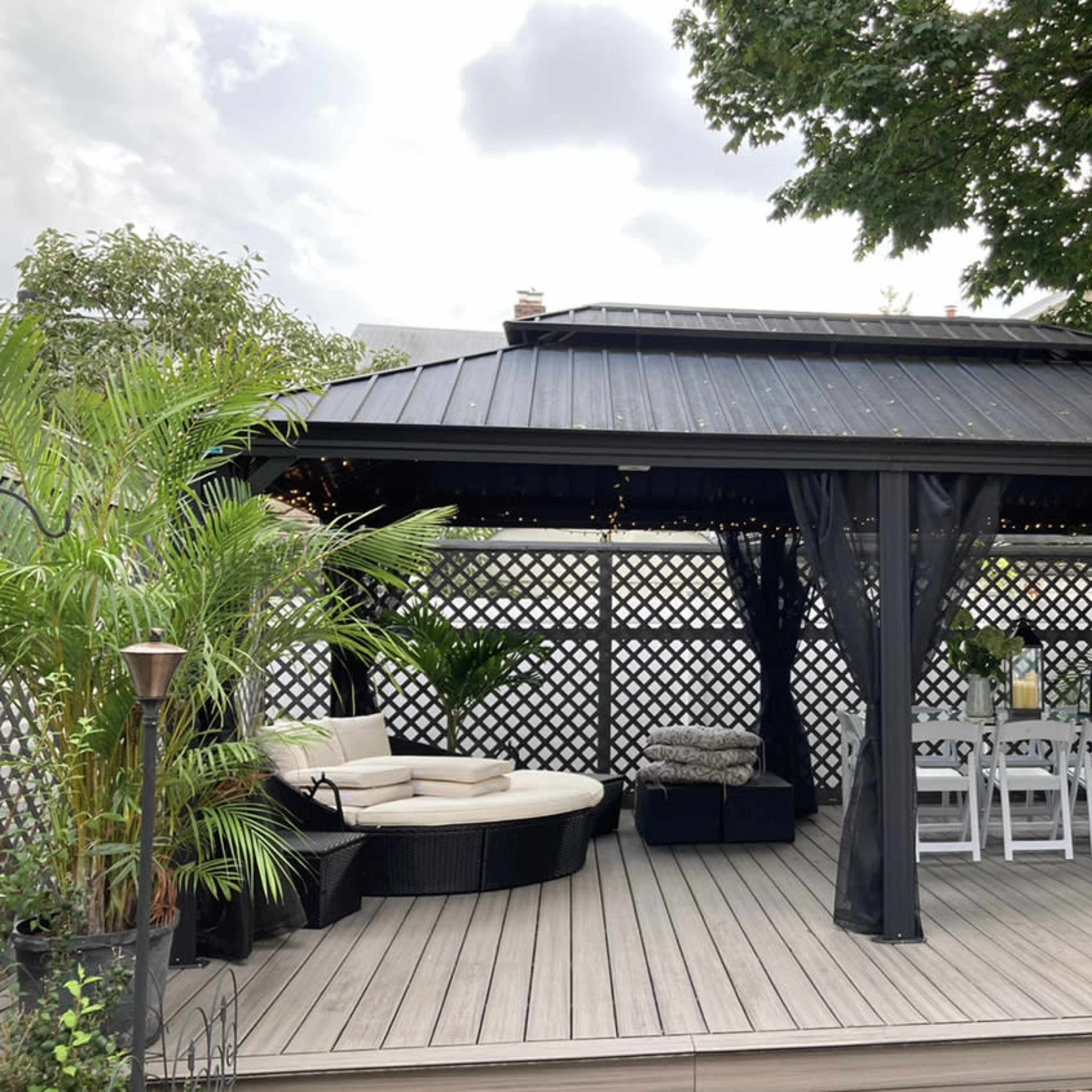 A black gazebo with a seating area and dining table, surrounded by greenery and decorative lights.