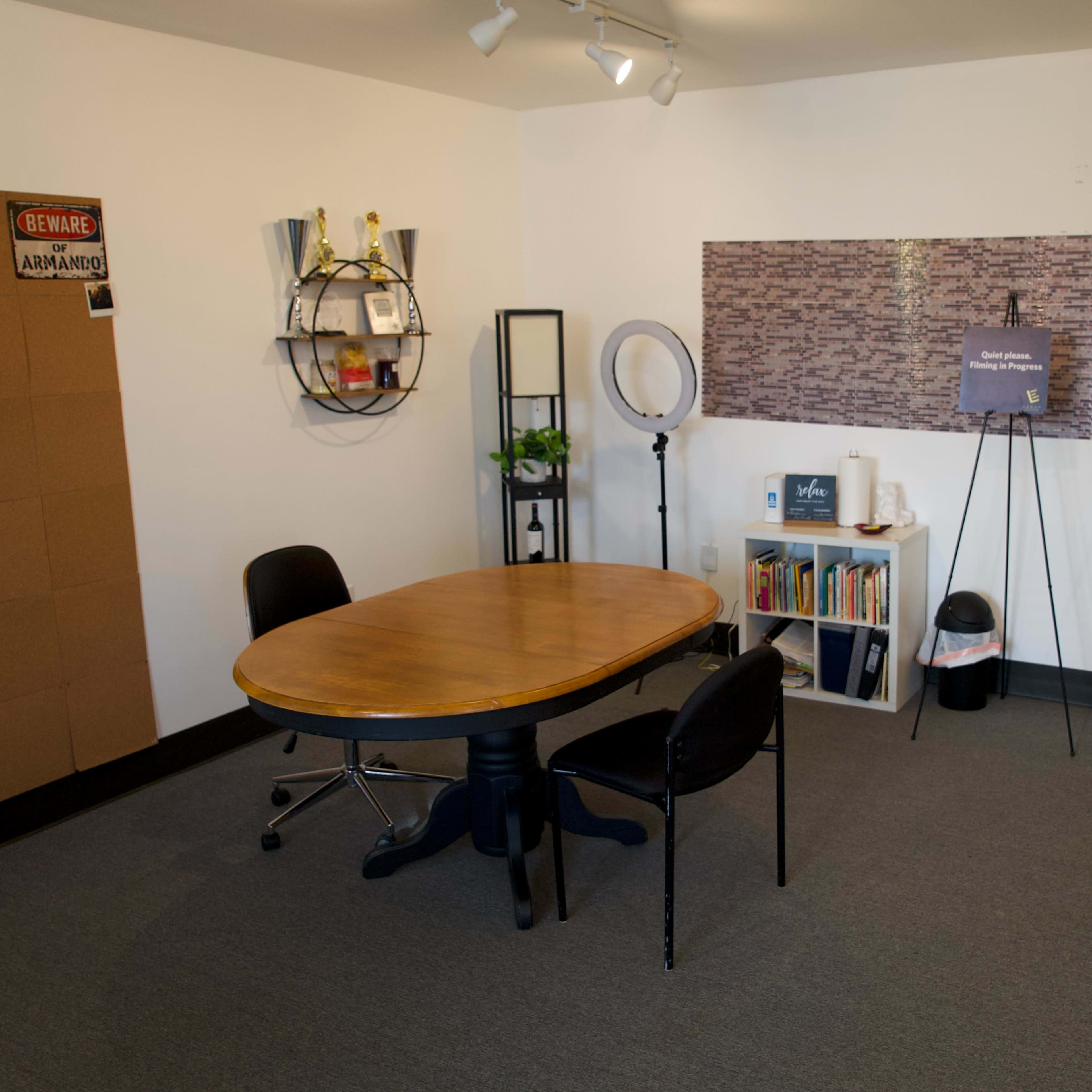 A minimalistic office space featuring a wooden round table with two black chairs, a bookshelf, a plant, and a board with pinned items on the wall.