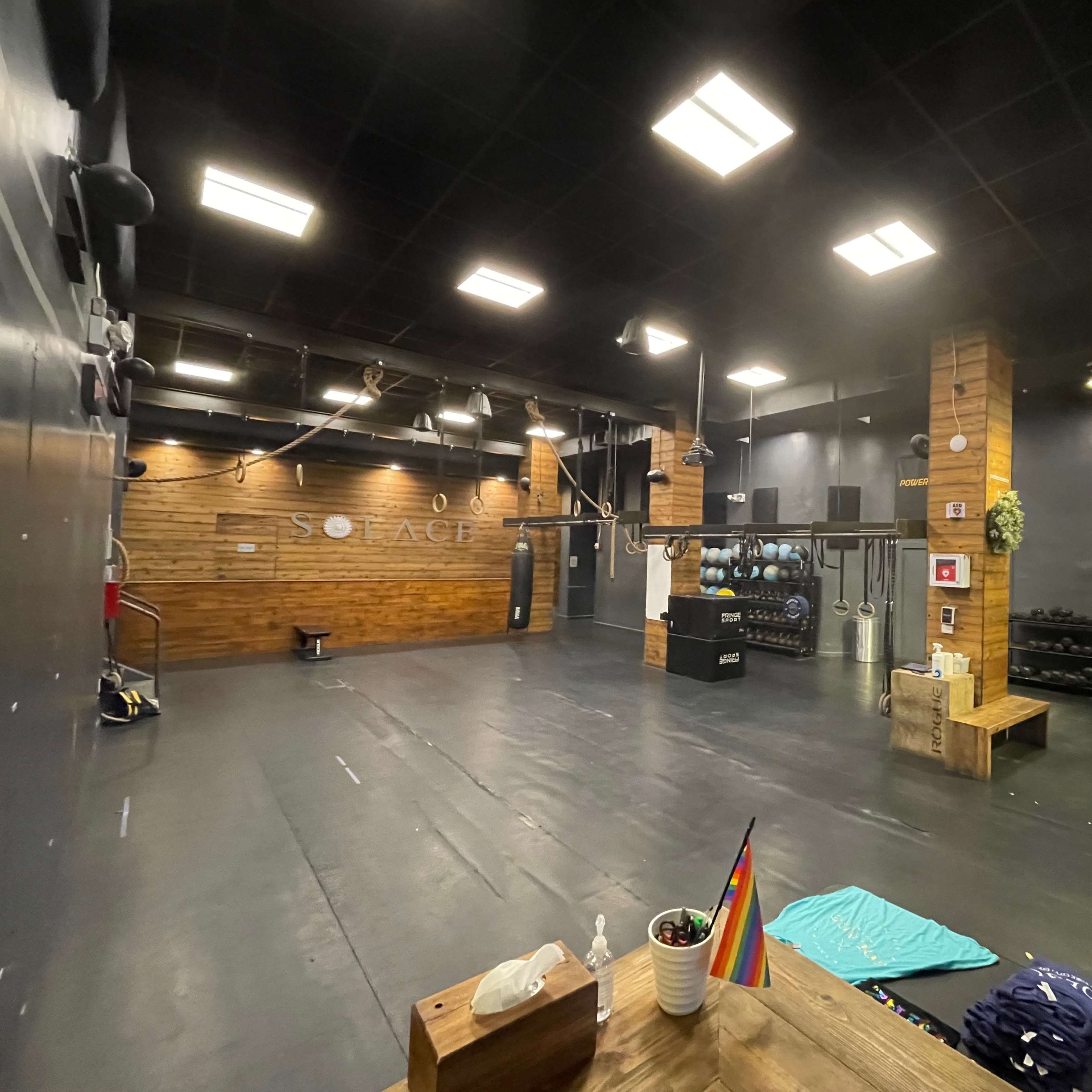 An industrial-style gym space with wooden accents, a row of rowing machines, and various fitness equipment arranged on a black floor under bright lights.