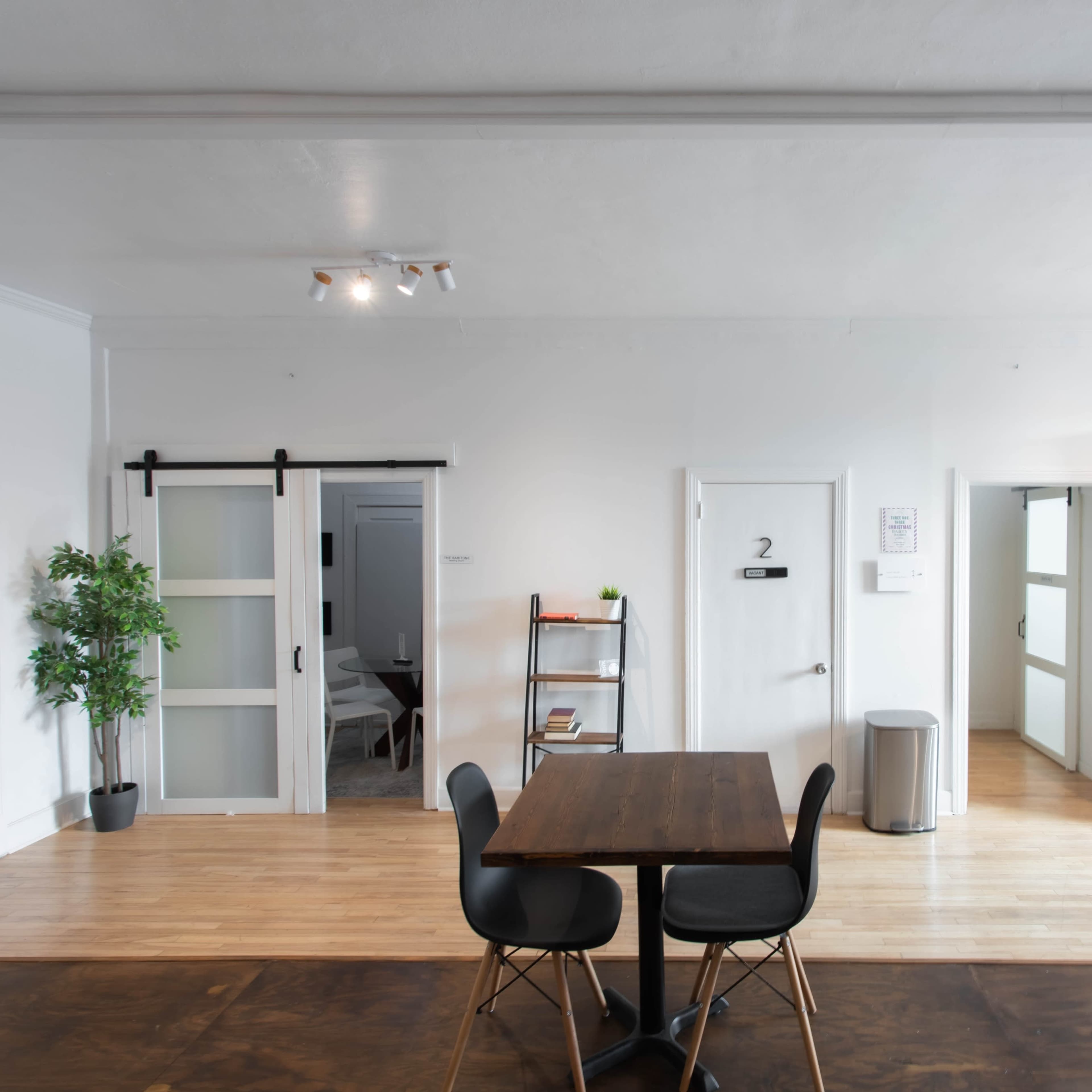A bright room features a wooden table with black chairs, a bookshelf filled with documents, and two doorways leading to separate areas.