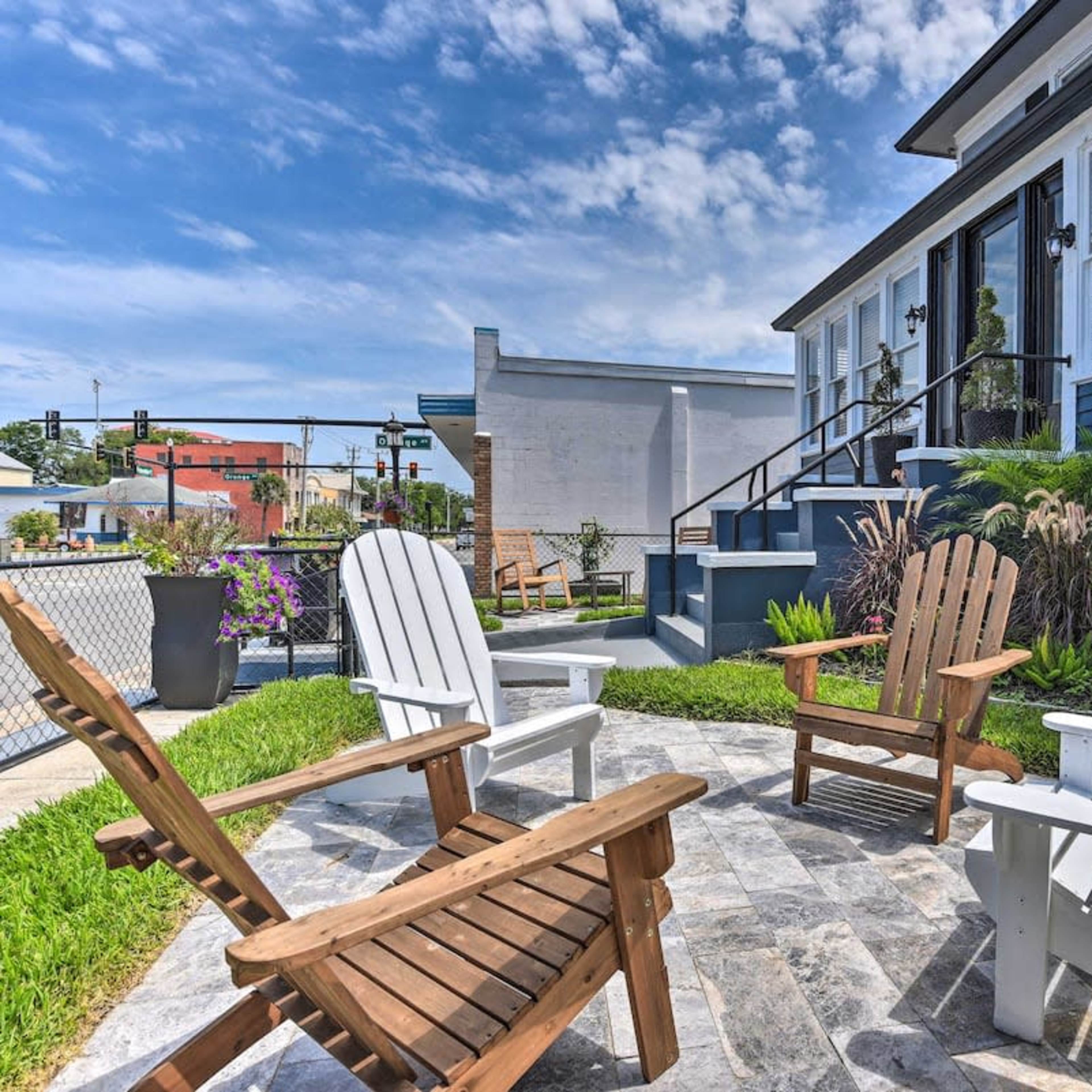 A cozy outdoor seating area with four Adirondack chairs arranged around a stone patio, near a neatly landscaped yard beside a house with blue siding.