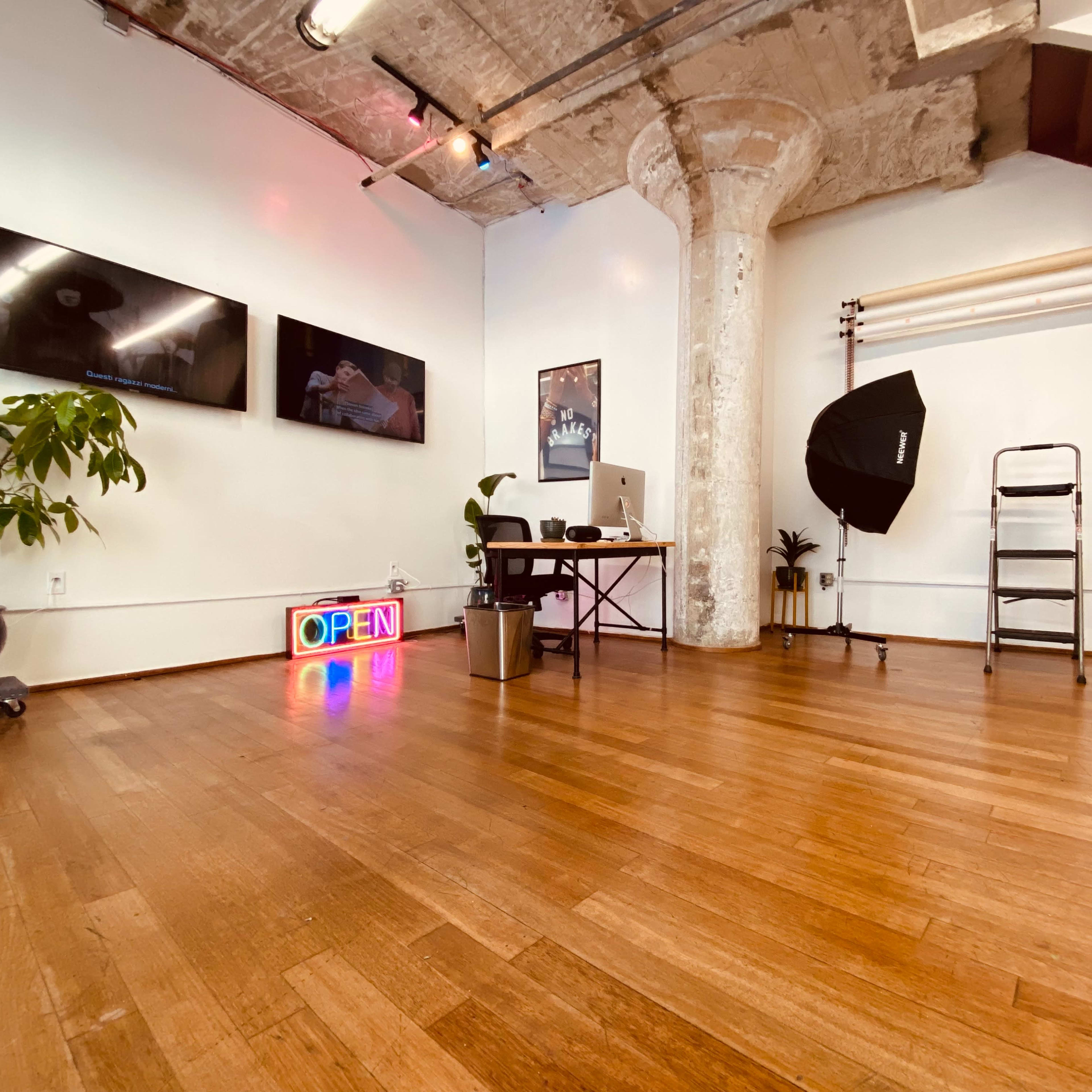 A bright, spacious studio workspace with hardwood floors, framed screens on the wall, and a neon "OPEN" sign, along with a plant and various photography equipment.