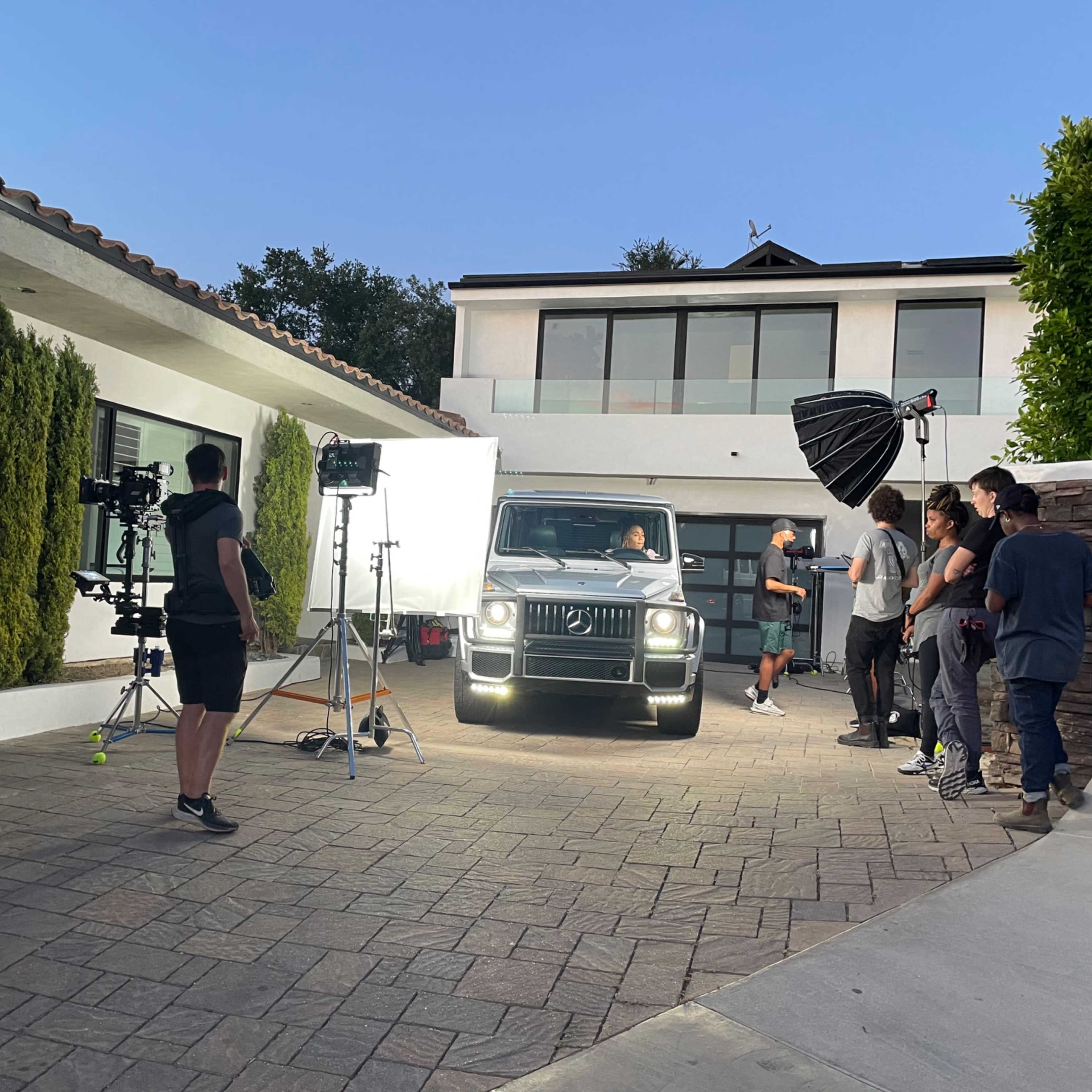 A film crew sets up lights and cameras while filming a Mercedes-Benz SUV outside a modern house.
