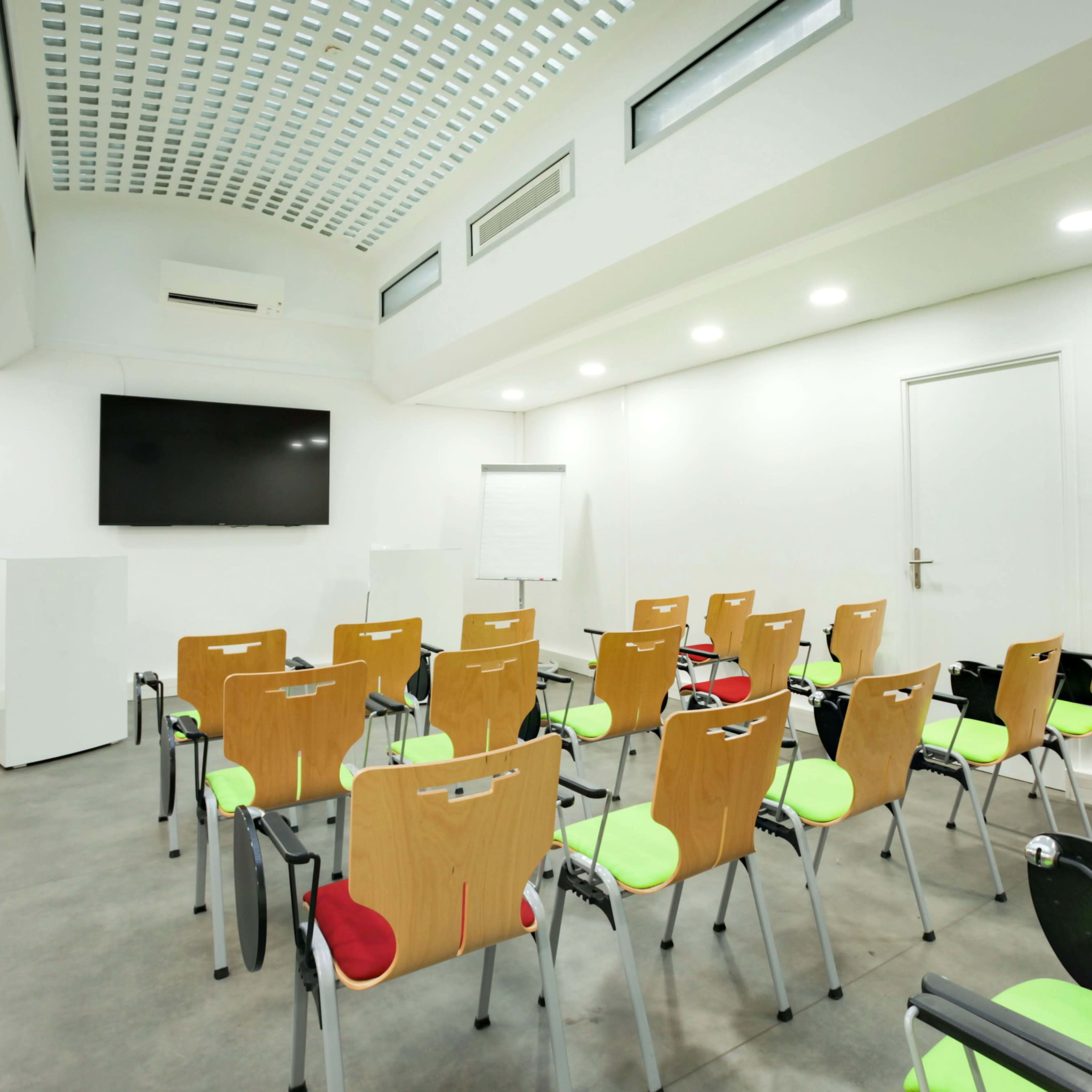 A modern meeting room with rows of wooden chairs featuring green and red seats, a large screen at the front, and a whiteboard.