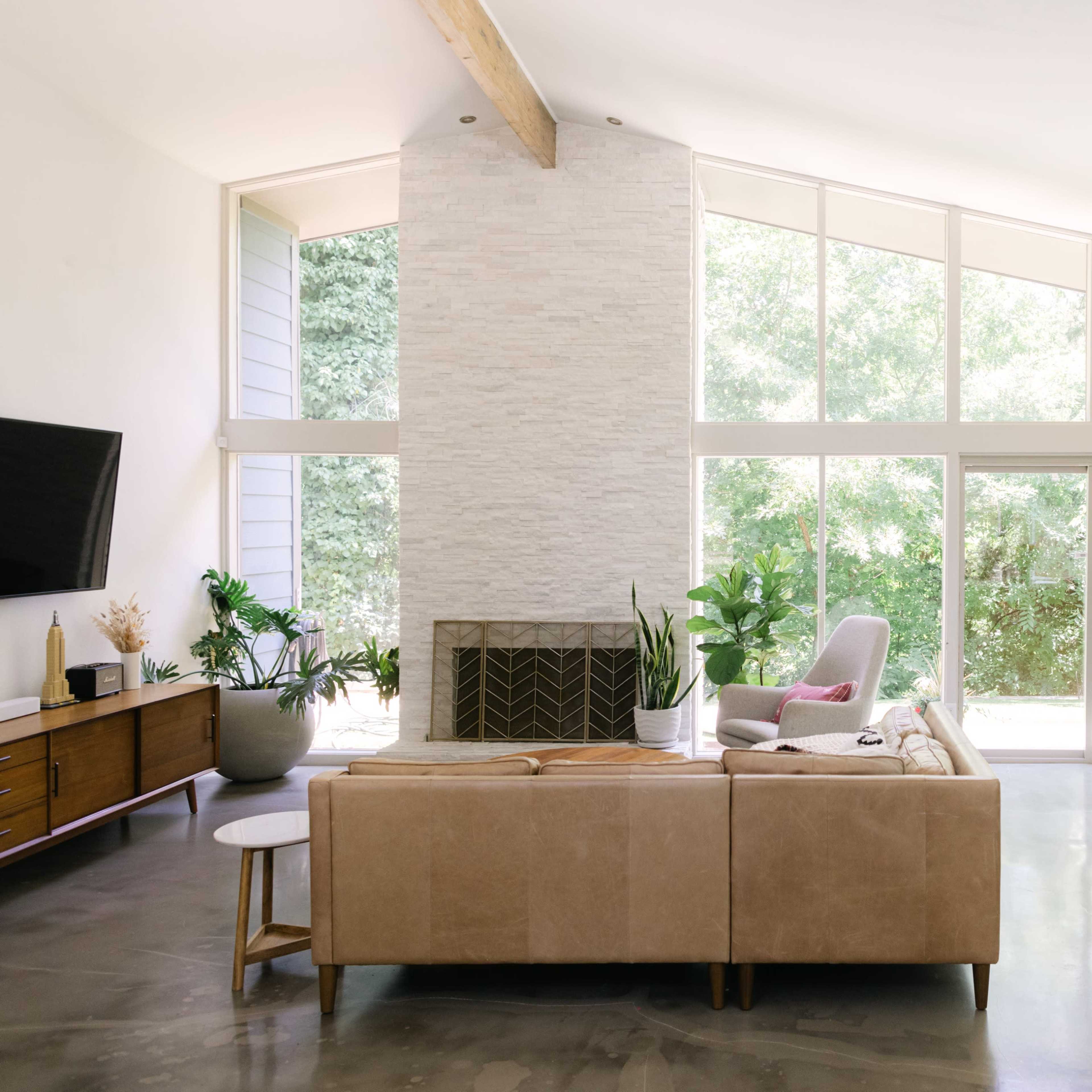 A bright living room with large windows overlooking a green landscape, featuring a beige sofa, a stone fireplace, and a dining area.