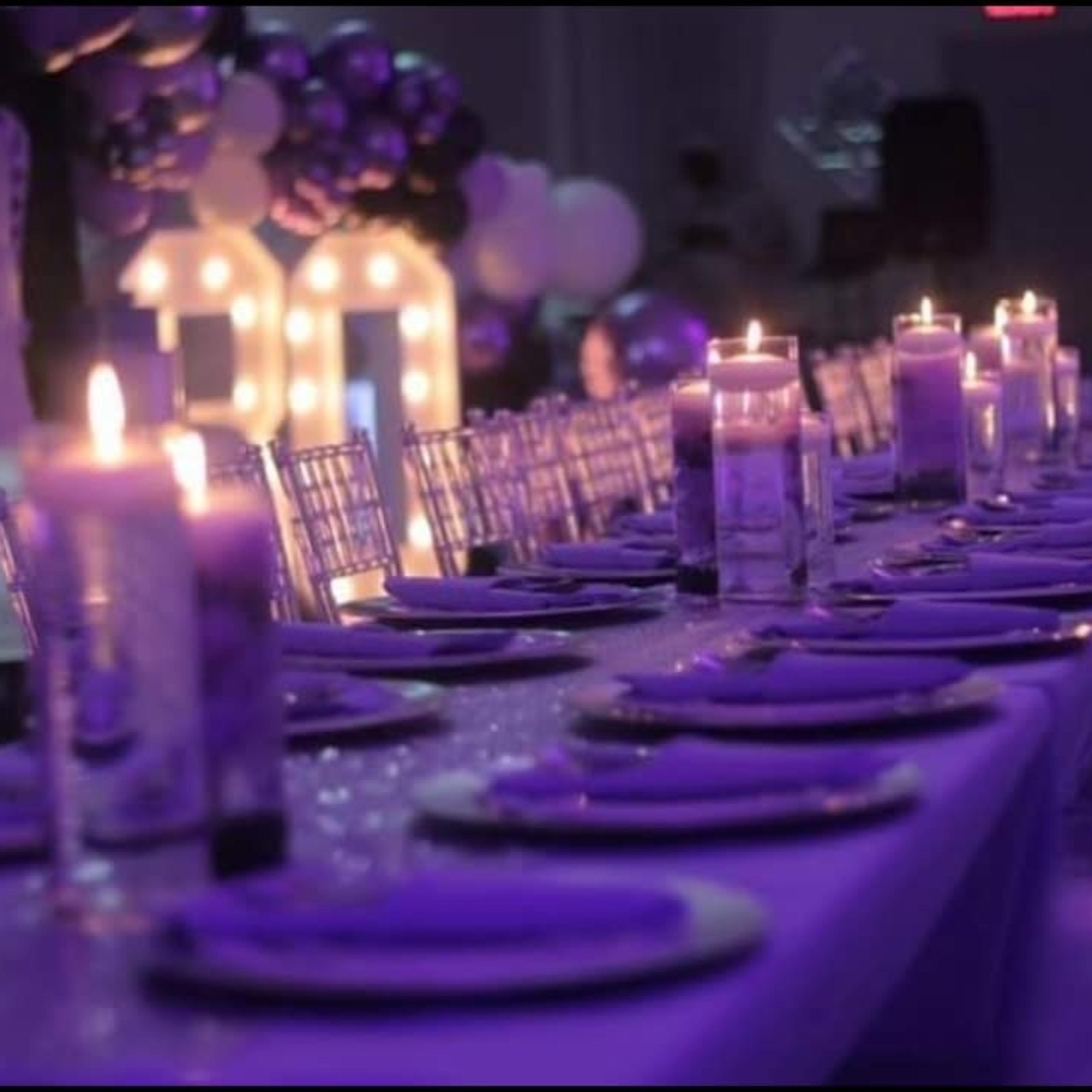 A long table is set for an event, featuring purple decorations, lit candles, and elegant place settings.