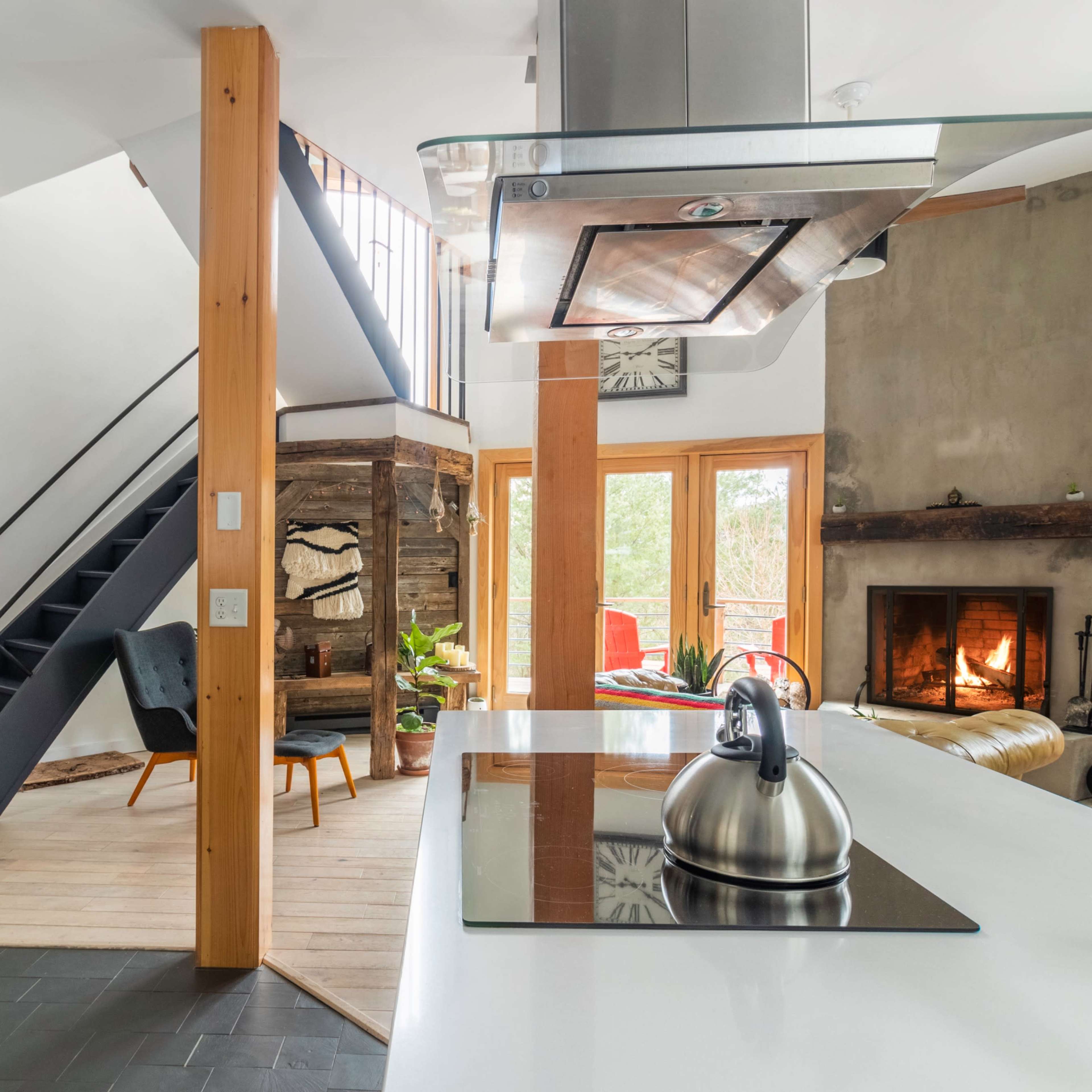 A modern kitchen with a stainless steel kettle on a white countertop, overlooking a staircase and a living area with a fireplace and seating.