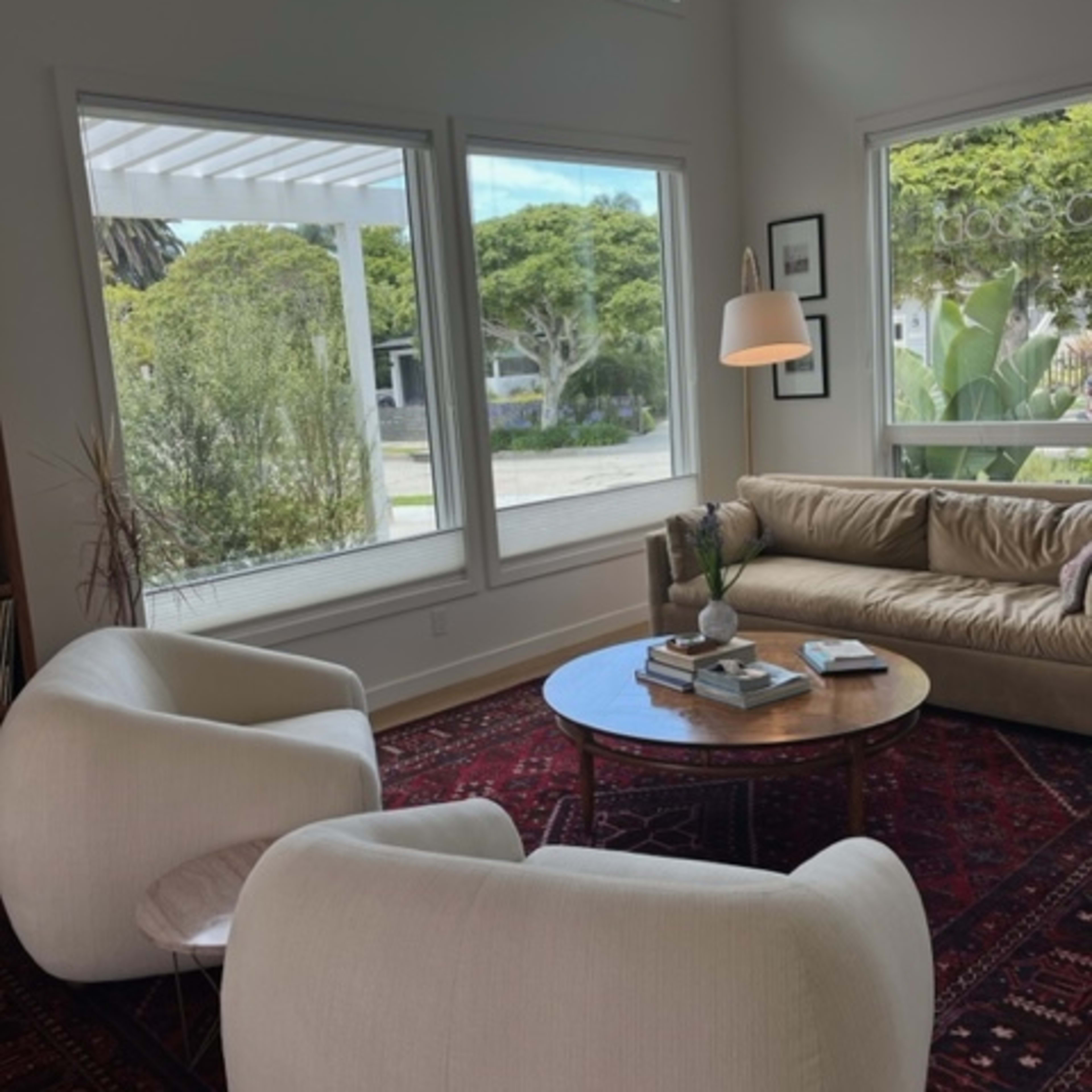A bright living room with two cream-colored chairs, a beige couch, and a round coffee table, all arranged near large windows that overlook a green landscape.