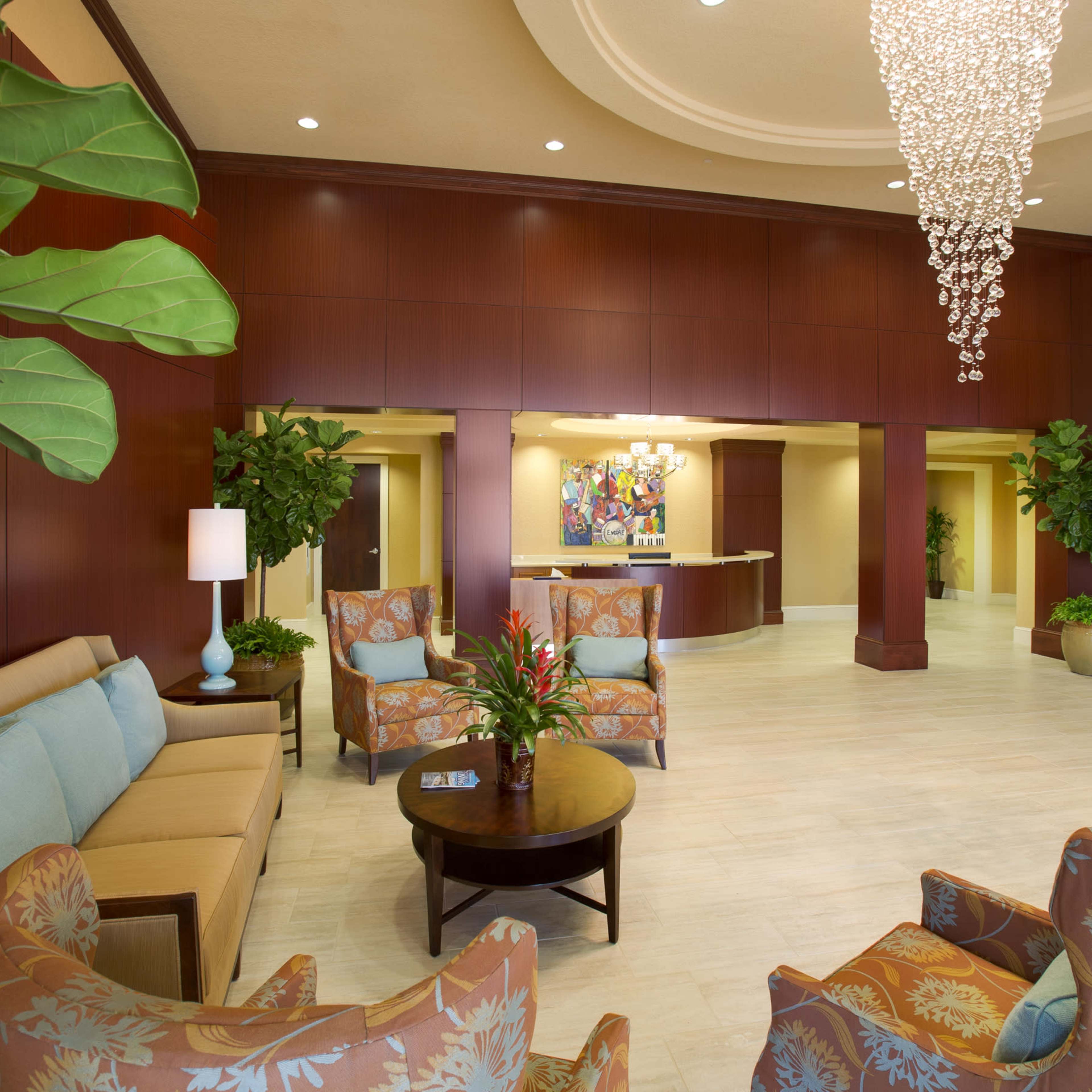 A brightly lit lobby area featuring a seating arrangement with patterned chairs, a coffee table, and decorative plants, alongside a reception desk in the background.