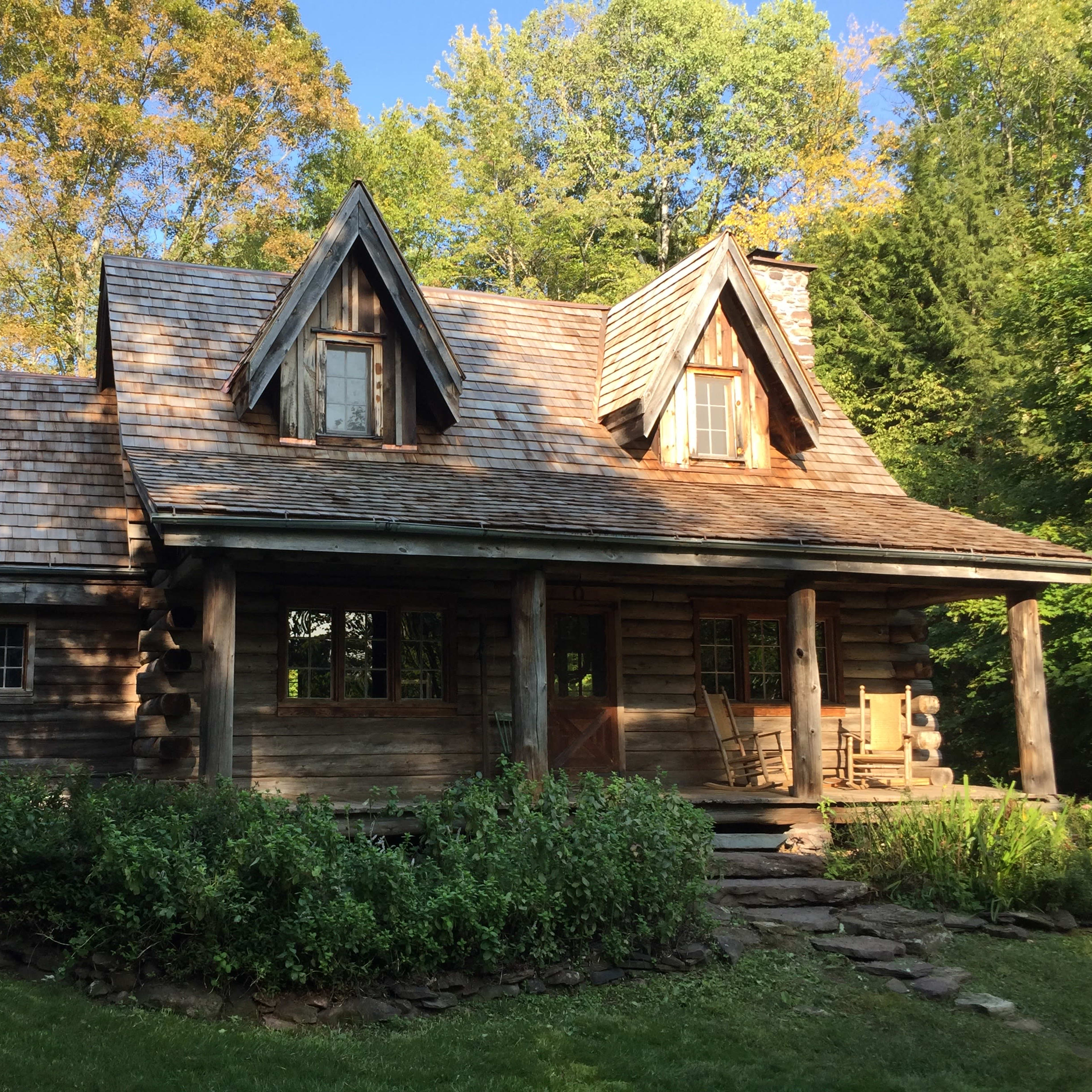 A wooden cabin with a steeply pitched roof and an inviting front porch is surrounded by lush greenery and trees.