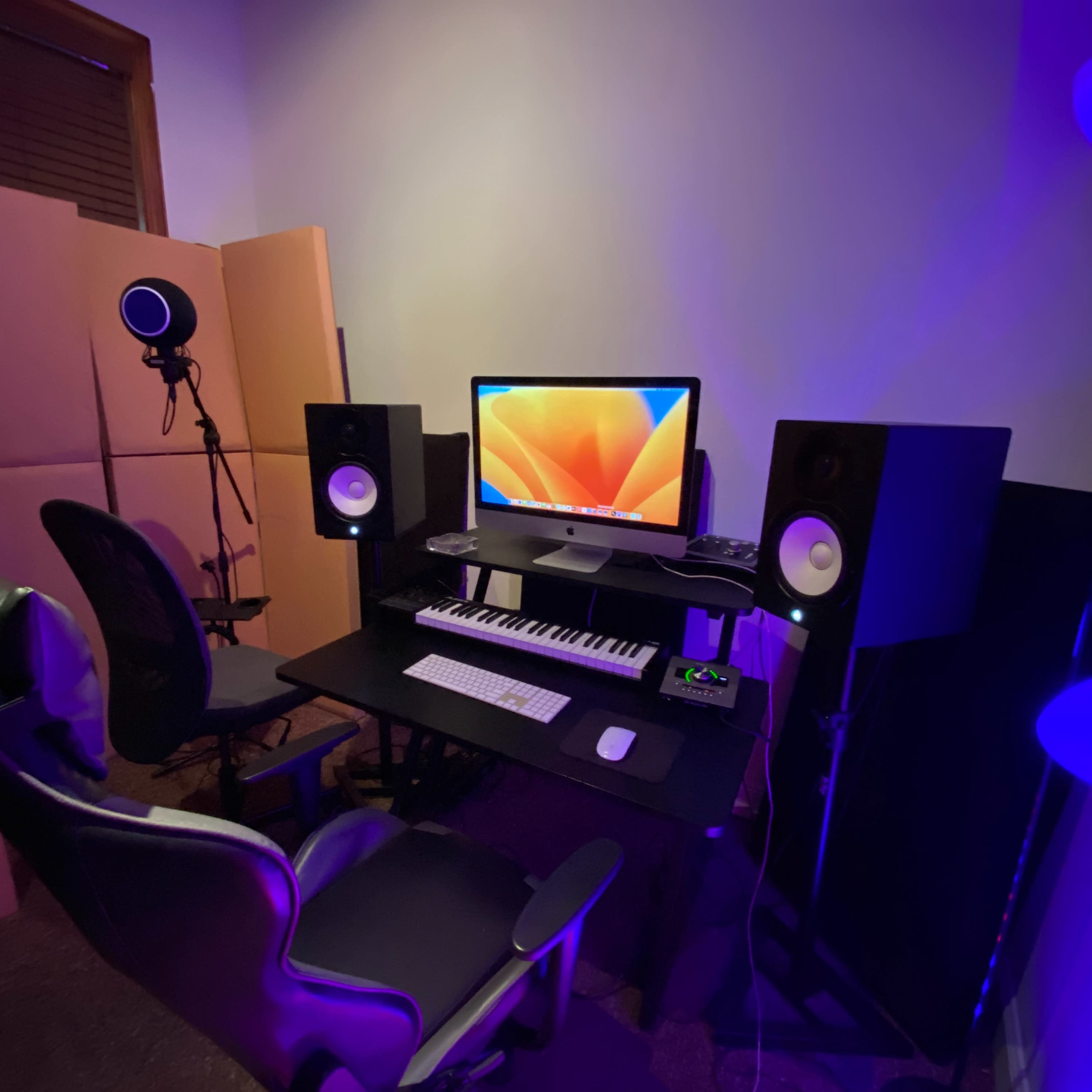 A music production setup featuring a computer, keyboard, two speakers, a microphone, and a chair, all situated in a dimly lit room.