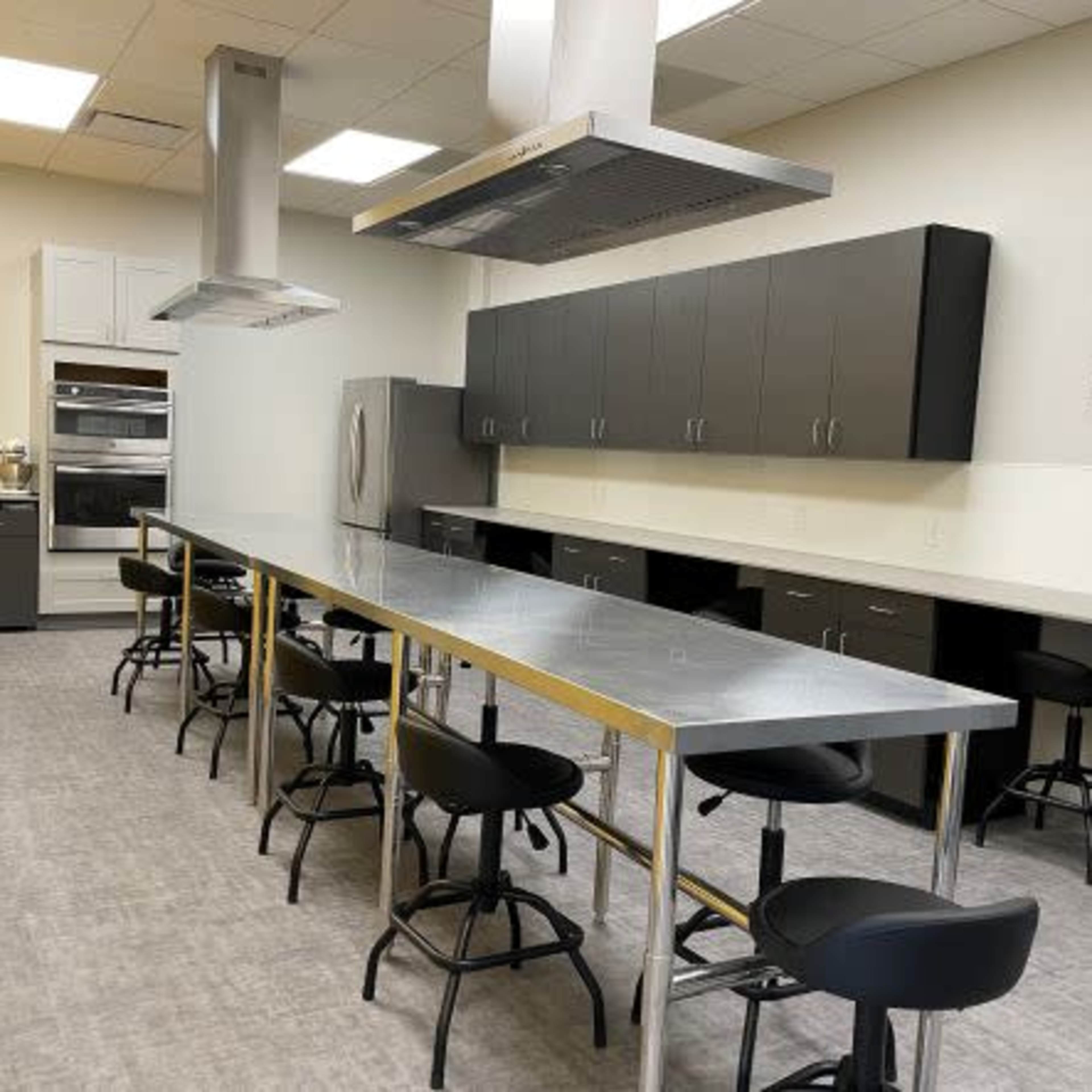 A modern kitchen features an elongated stainless steel countertop with barstools, dark cabinetry, and stainless steel appliances along one wall.