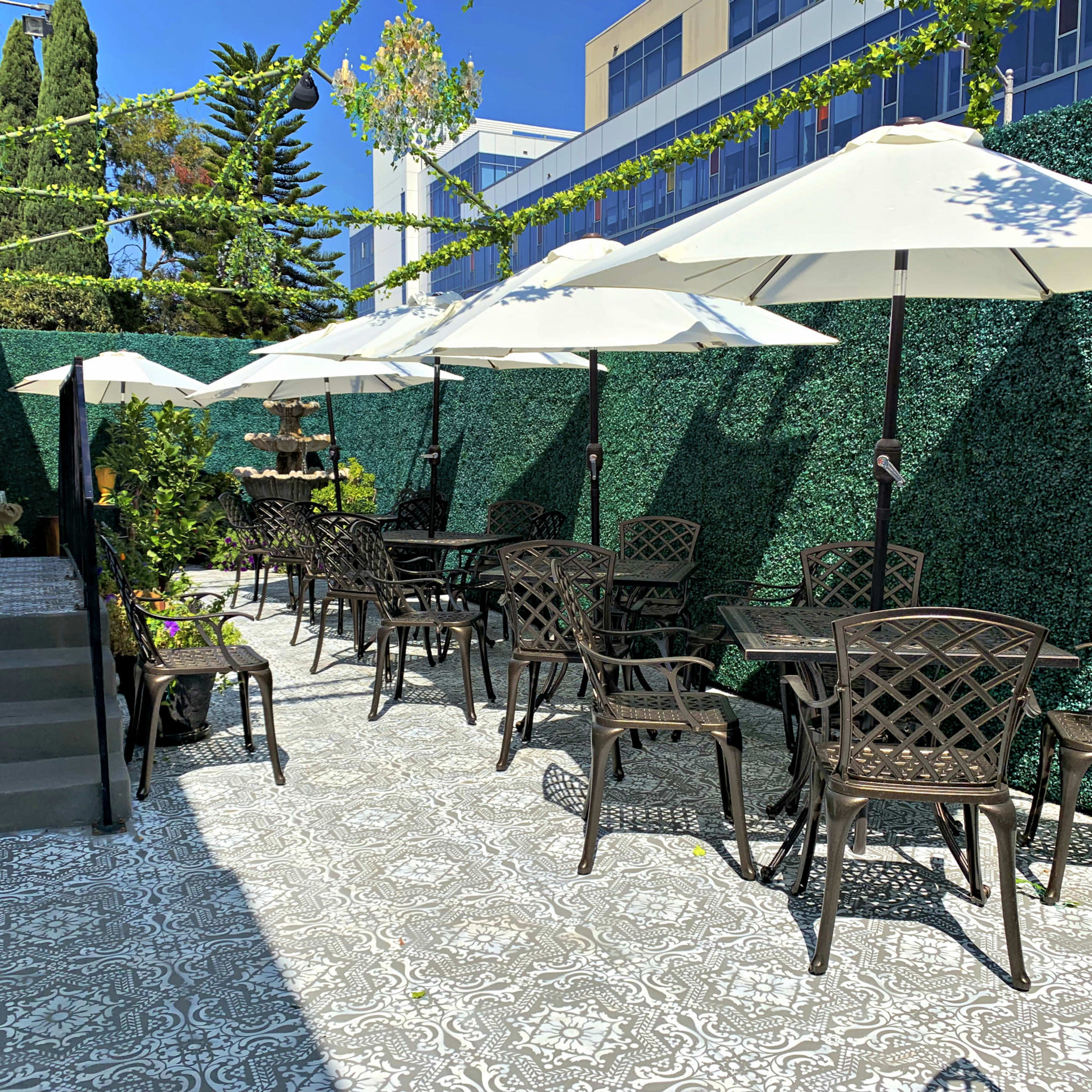A patio area with several tables and chairs under white umbrellas, surrounded by green hedges and stepping stones.