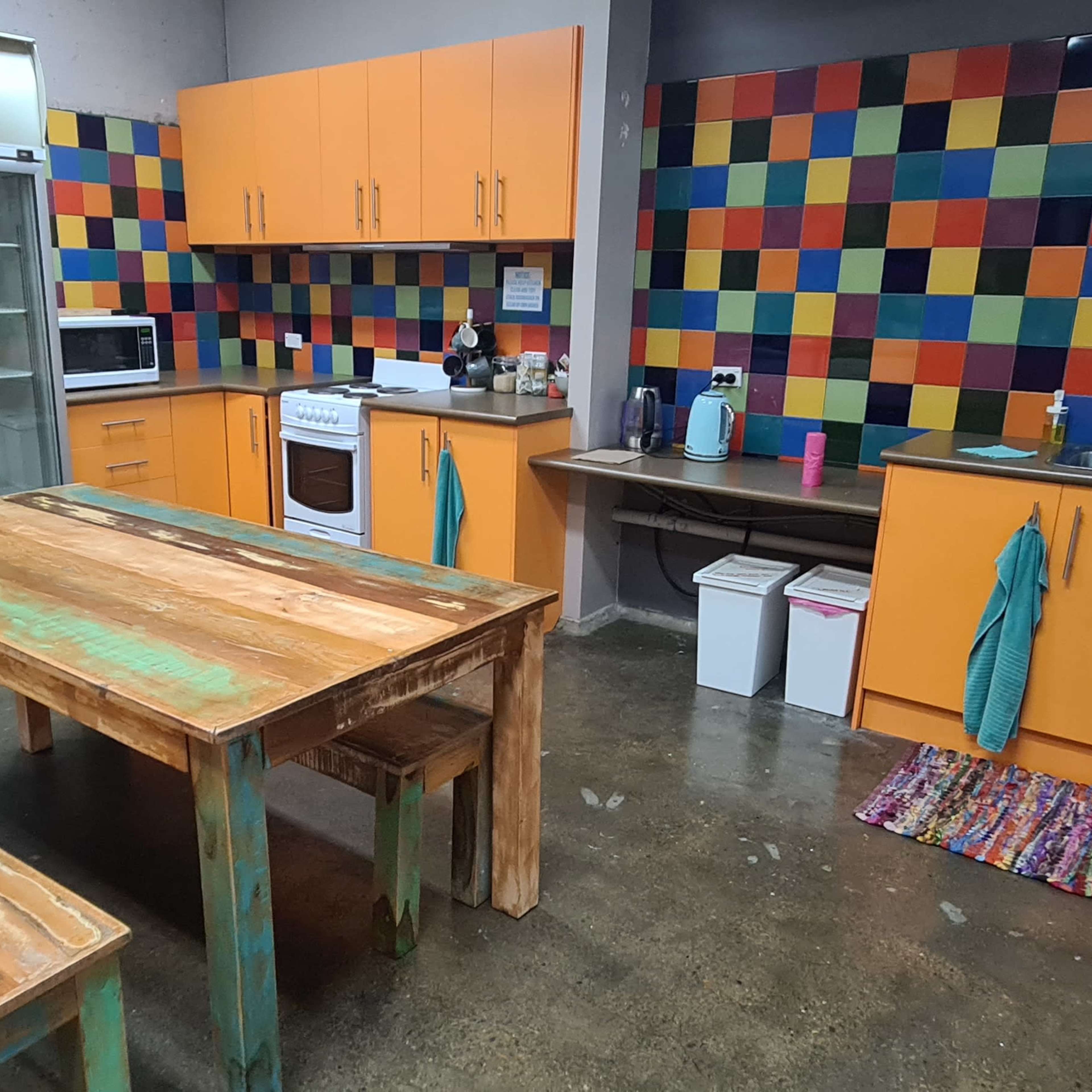 A kitchen with orange cabinets, a wooden table and benches, colorful tiled walls, and various appliances.