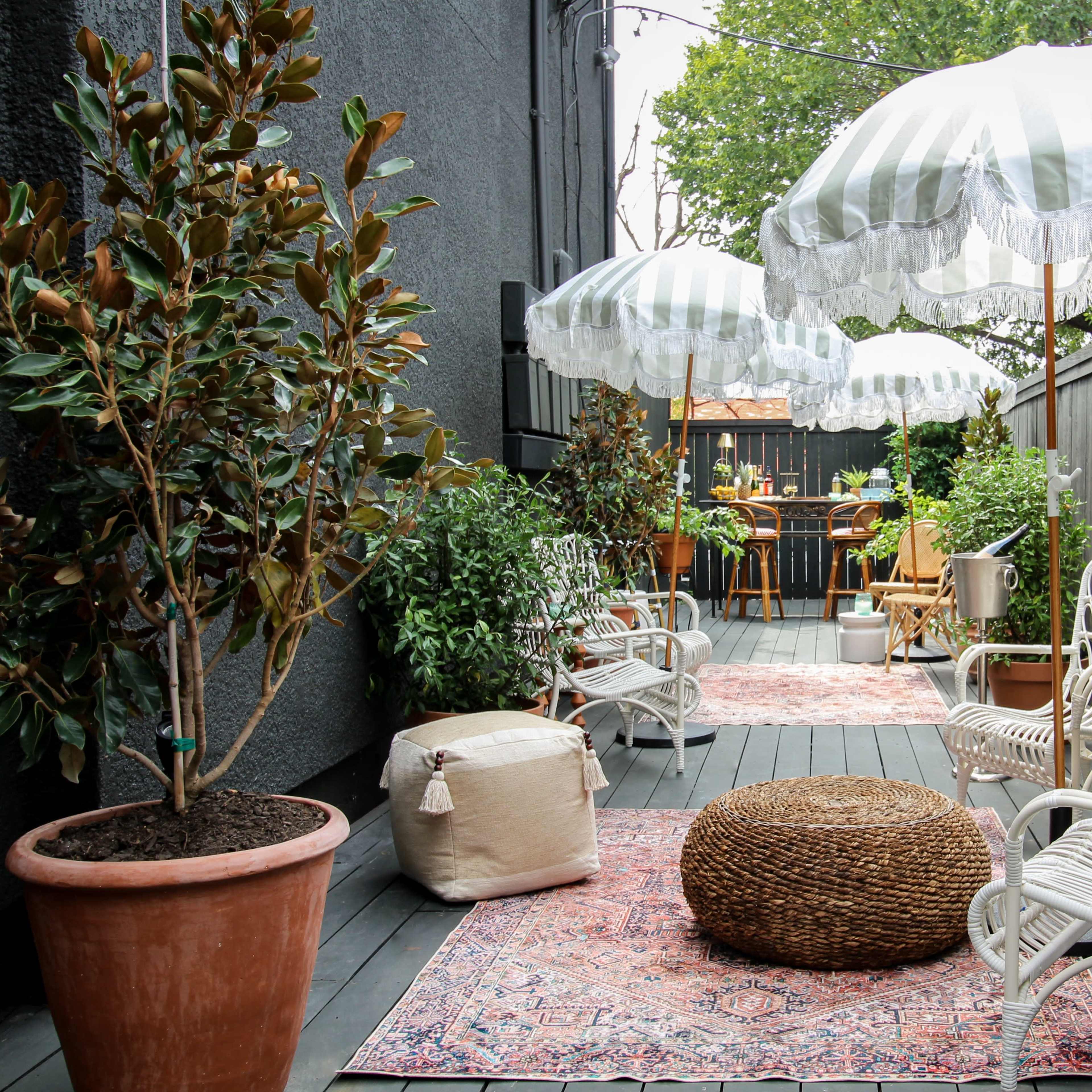 A narrow outdoor space featuring white wicker chairs, striped umbrellas, potted plants, and a patterned rug on wooden decking.