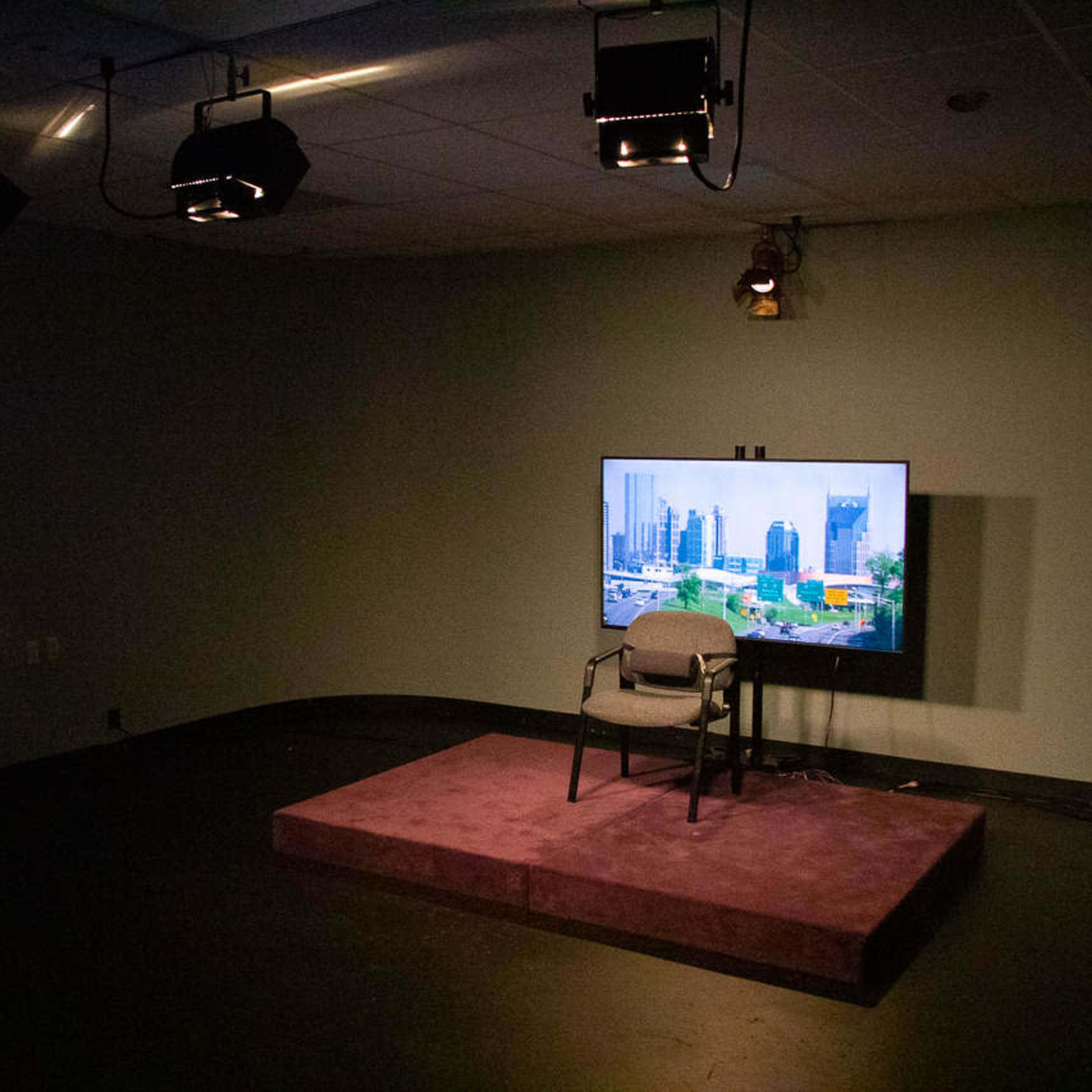 A dimly lit studio with a single chair on a purple stage, a television displaying a cityscape, and spotlights overhead.