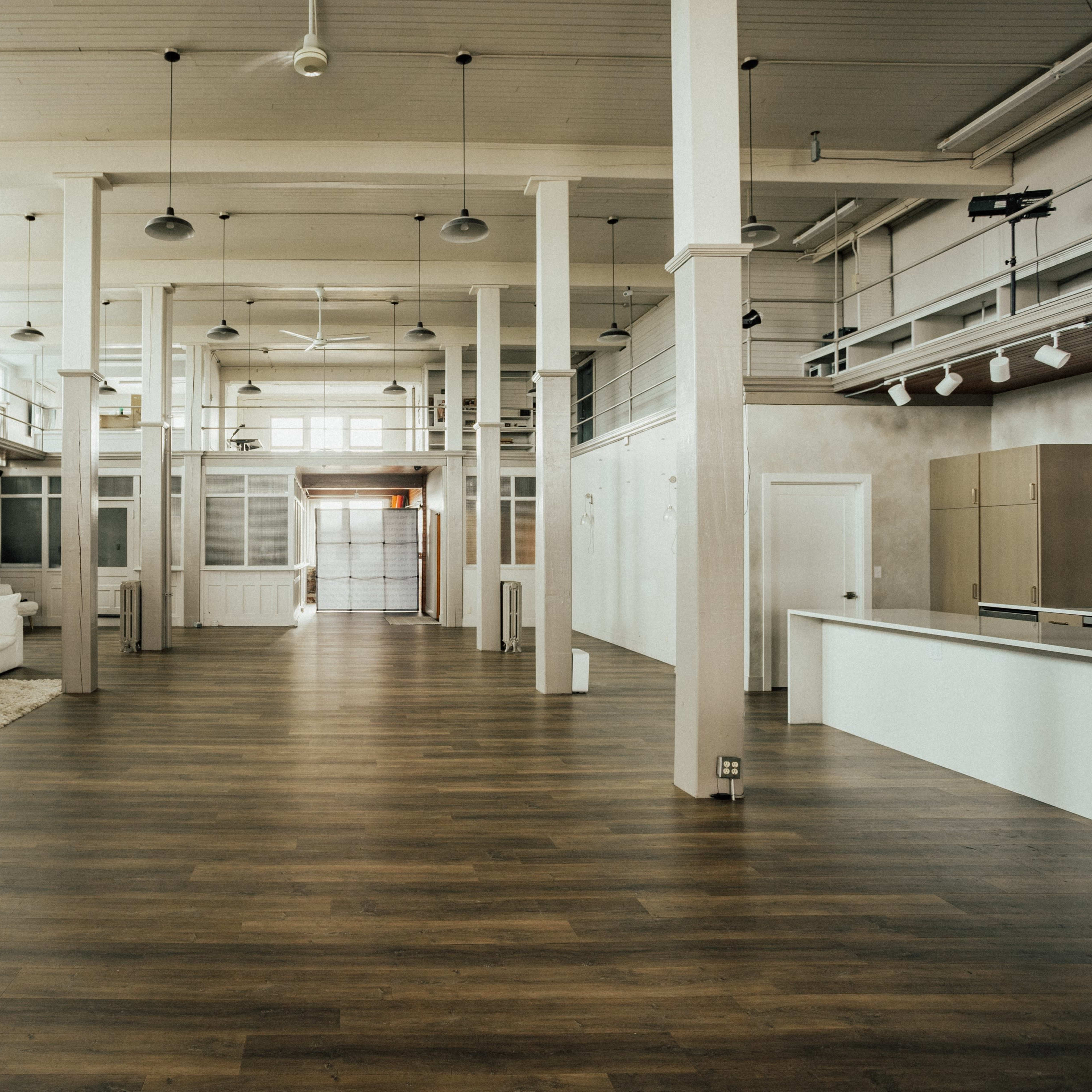 A spacious, modern interior with wooden floors, tall white columns, and large windows, featuring minimal furnishings and an open layout.