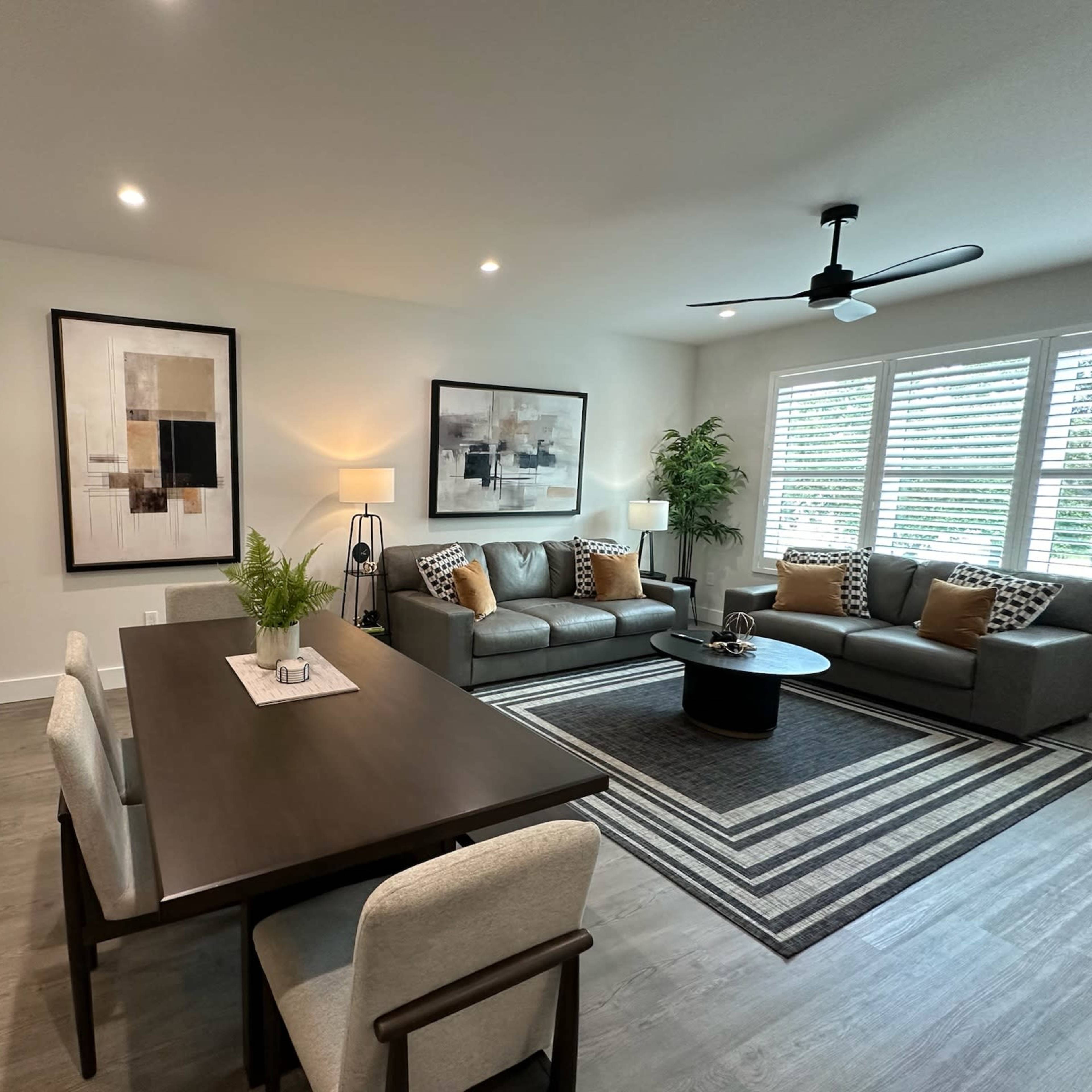 A modern living room and dining area featuring a gray sofa set, a round coffee table, and a dining table with chairs, all set against a backdrop of large windows with blinds.