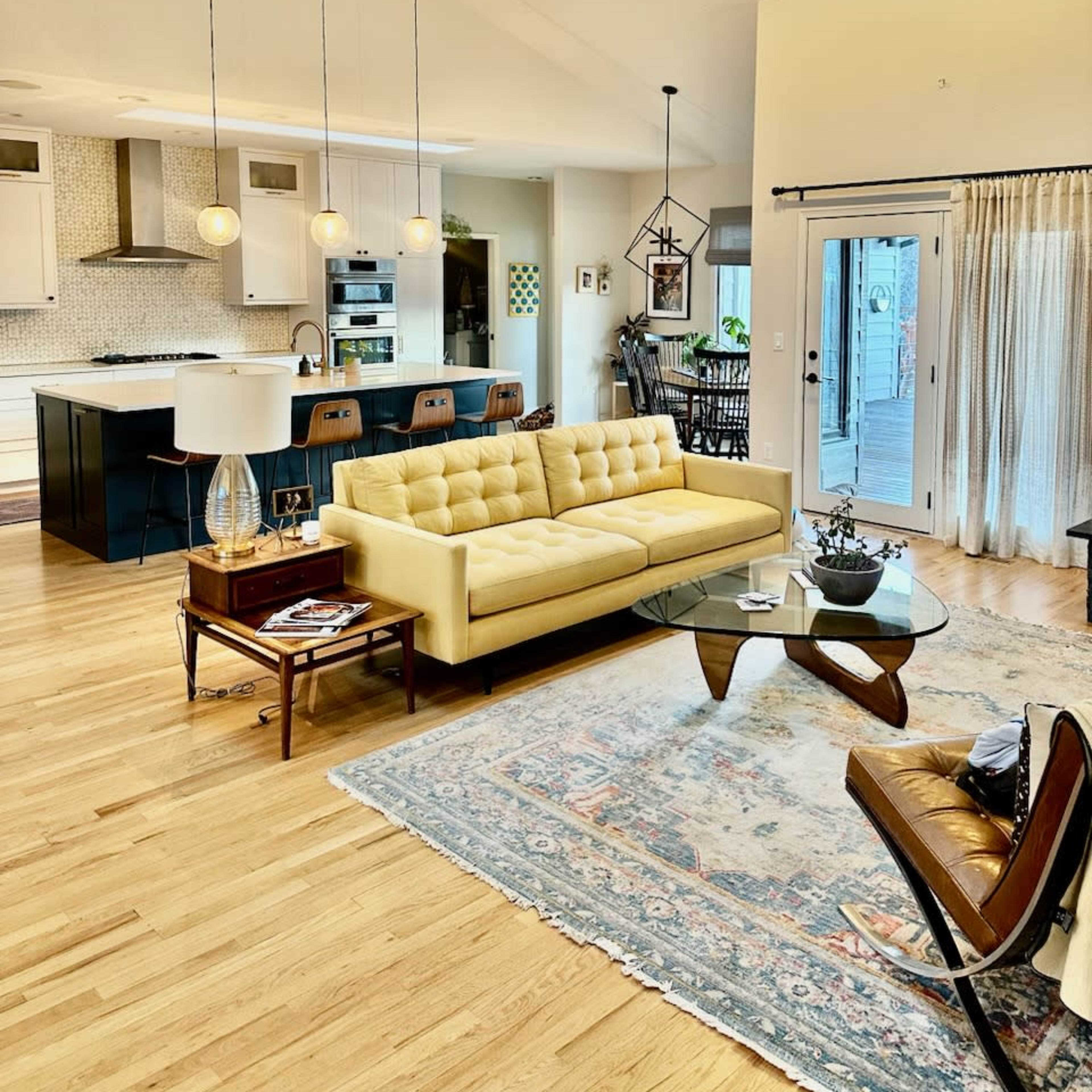 A bright living area features a yellow tufted sofa, a glass coffee table, and a modern kitchen visible in the background.