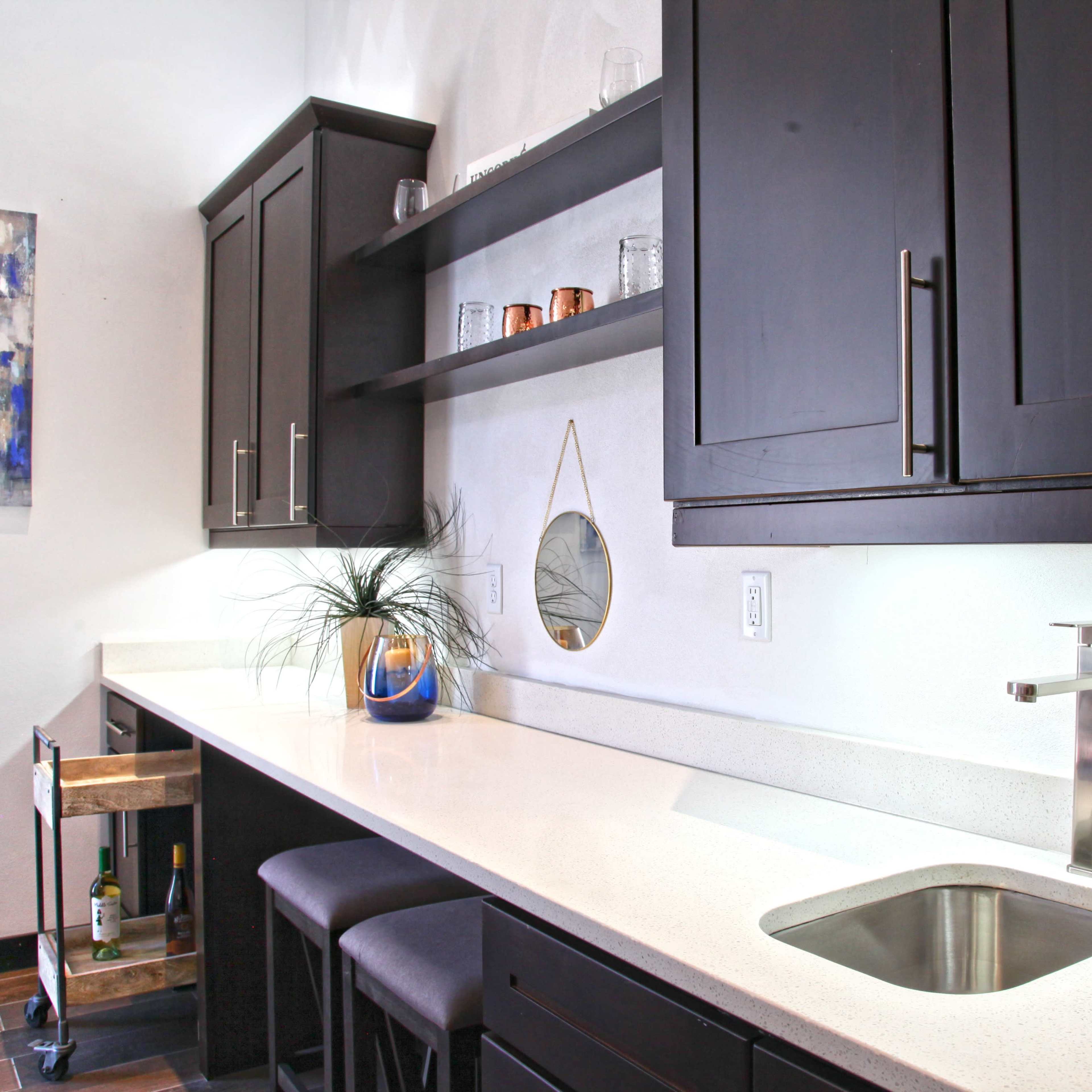 A modern kitchen features dark cabinetry, a sleek countertop, an undermount sink, and a small cart with beverages.