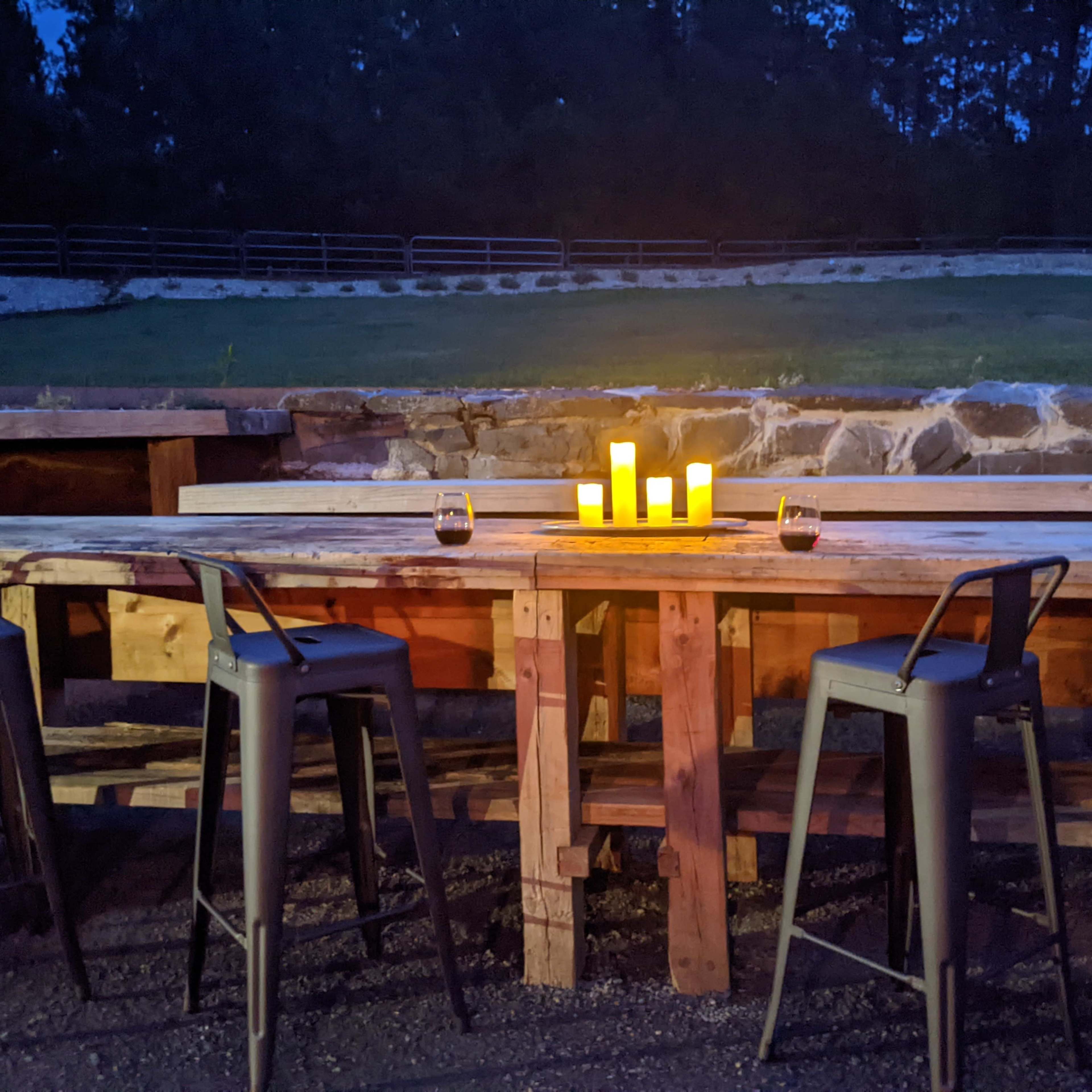 A wooden bar table with high stools is illuminated by candles in a dimly lit outdoor setting.