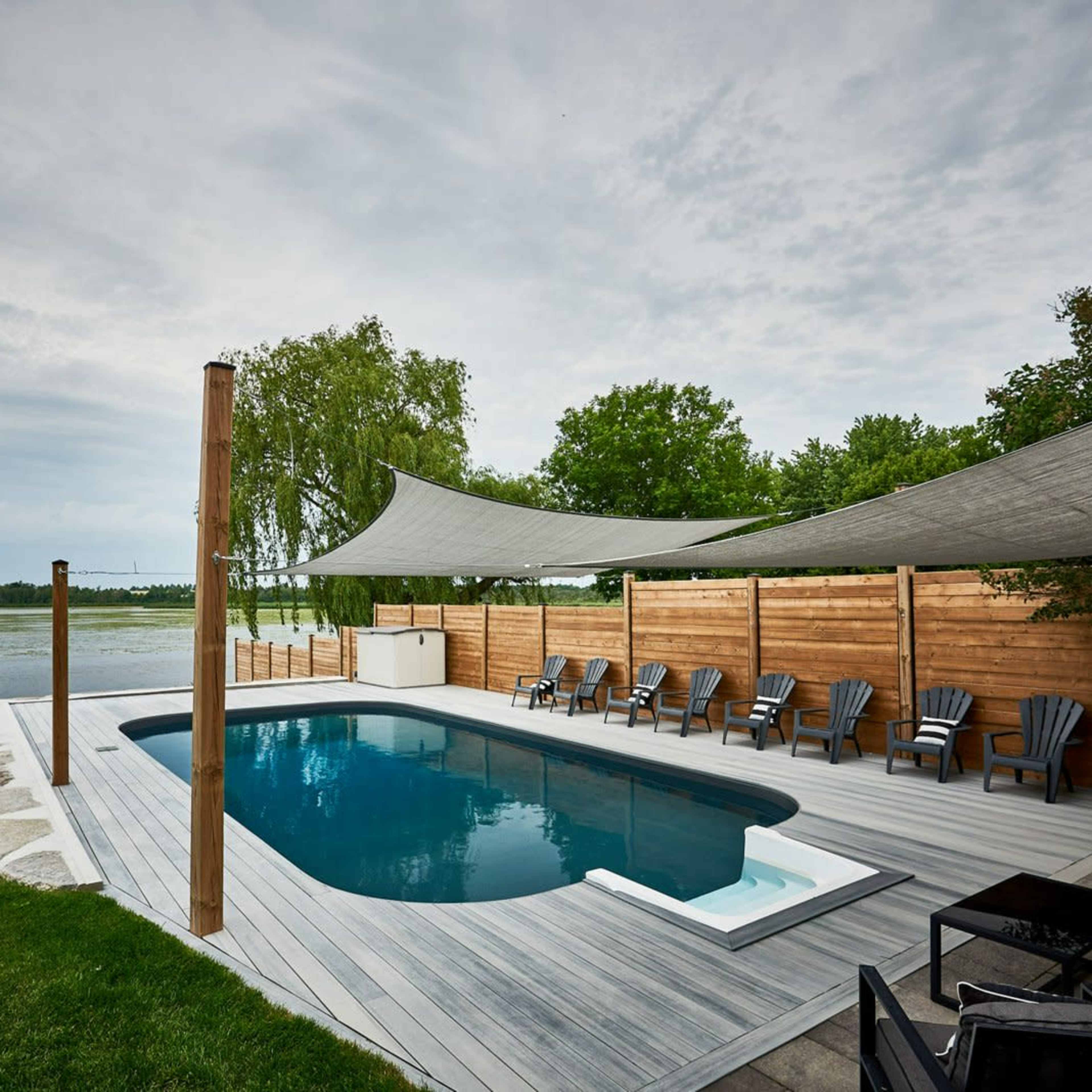 A swimming pool surrounded by wooden decking and chairs, with shade sails overhead, next to a calm body of water and green trees.