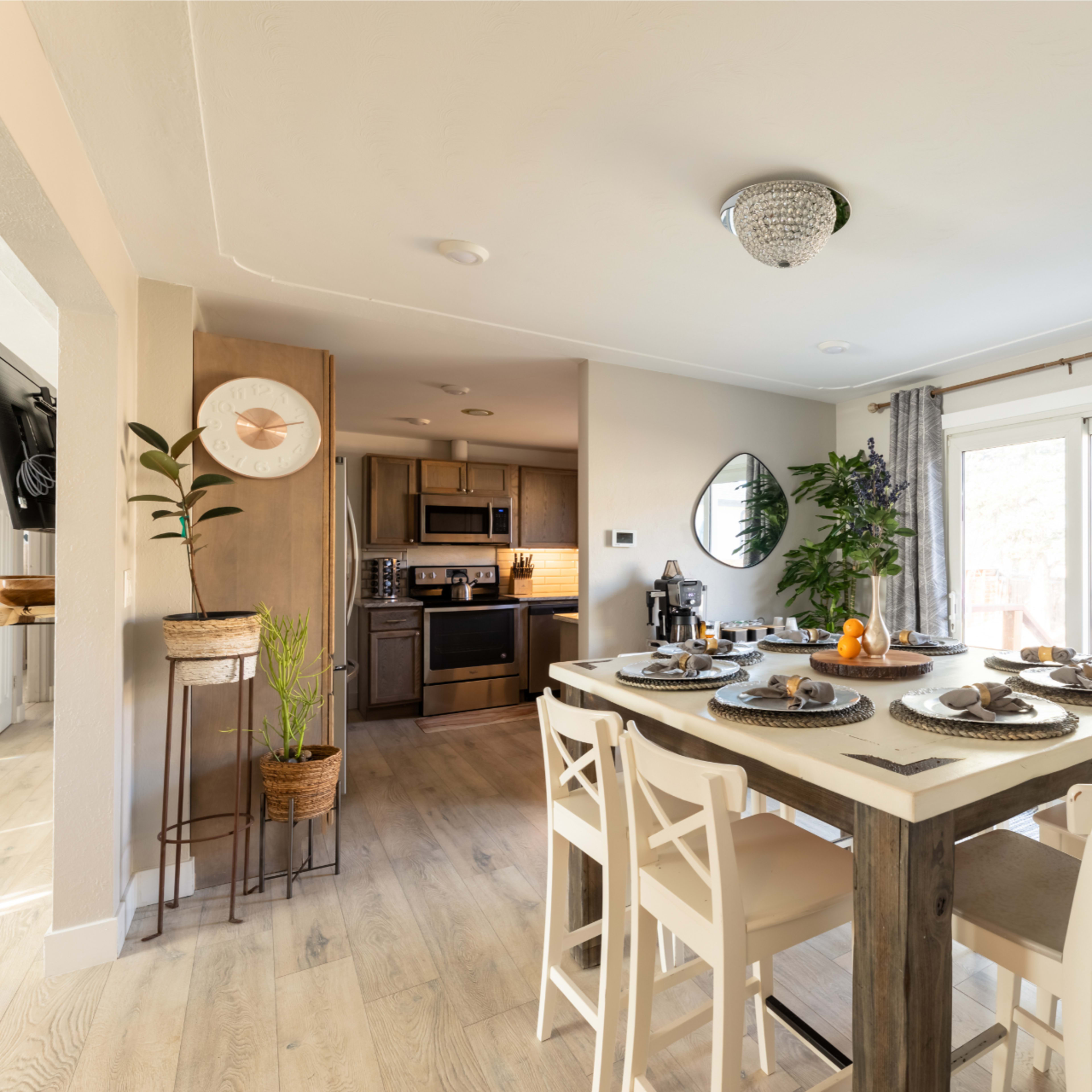 A modern dining area features a wooden table set for a meal, with an adjacent kitchen visible in the background and large windows allowing natural light to enter.