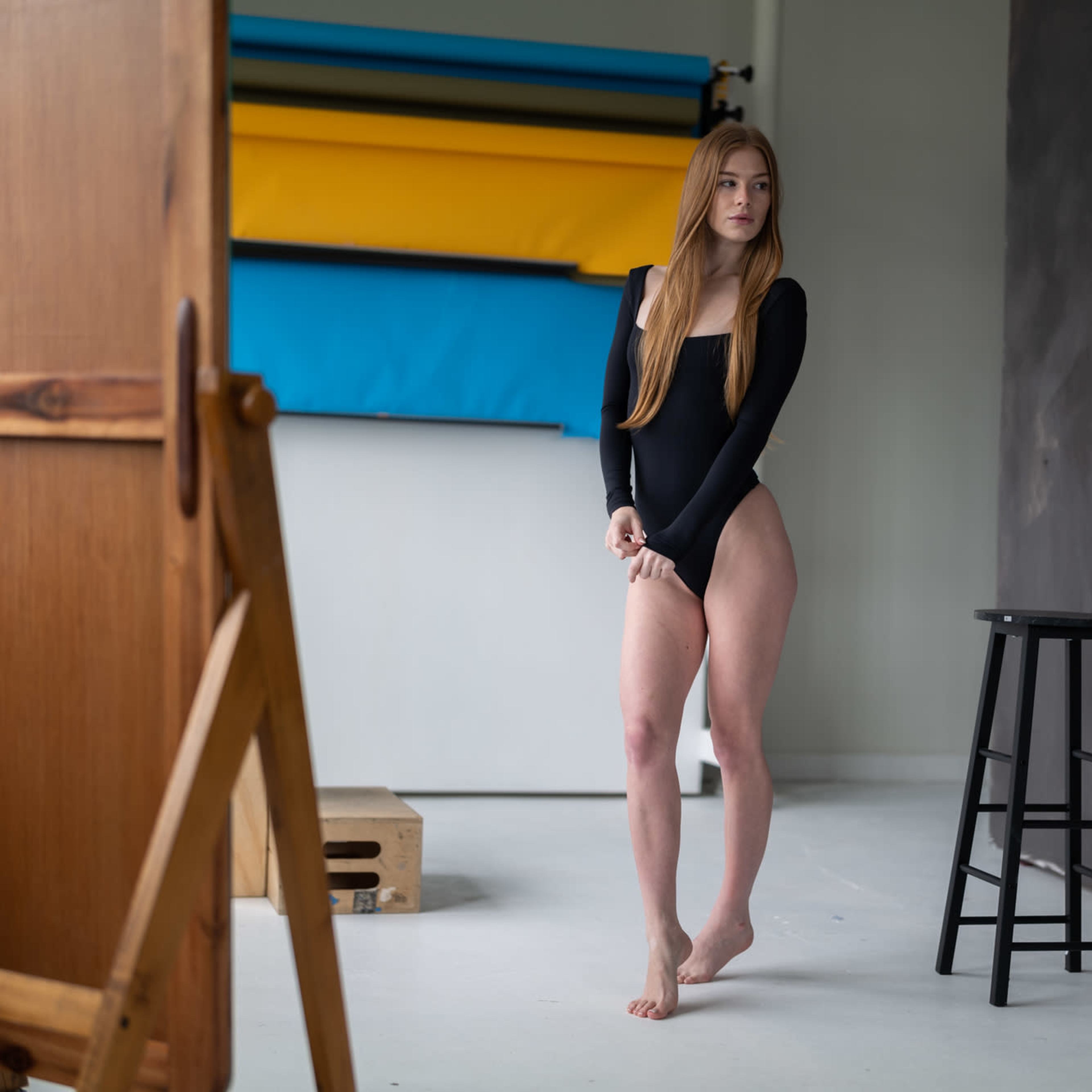 A model stands beside an easel in a photography studio, contrasting a black bodysuit with the vibrant backdrop of colored paper rolls.