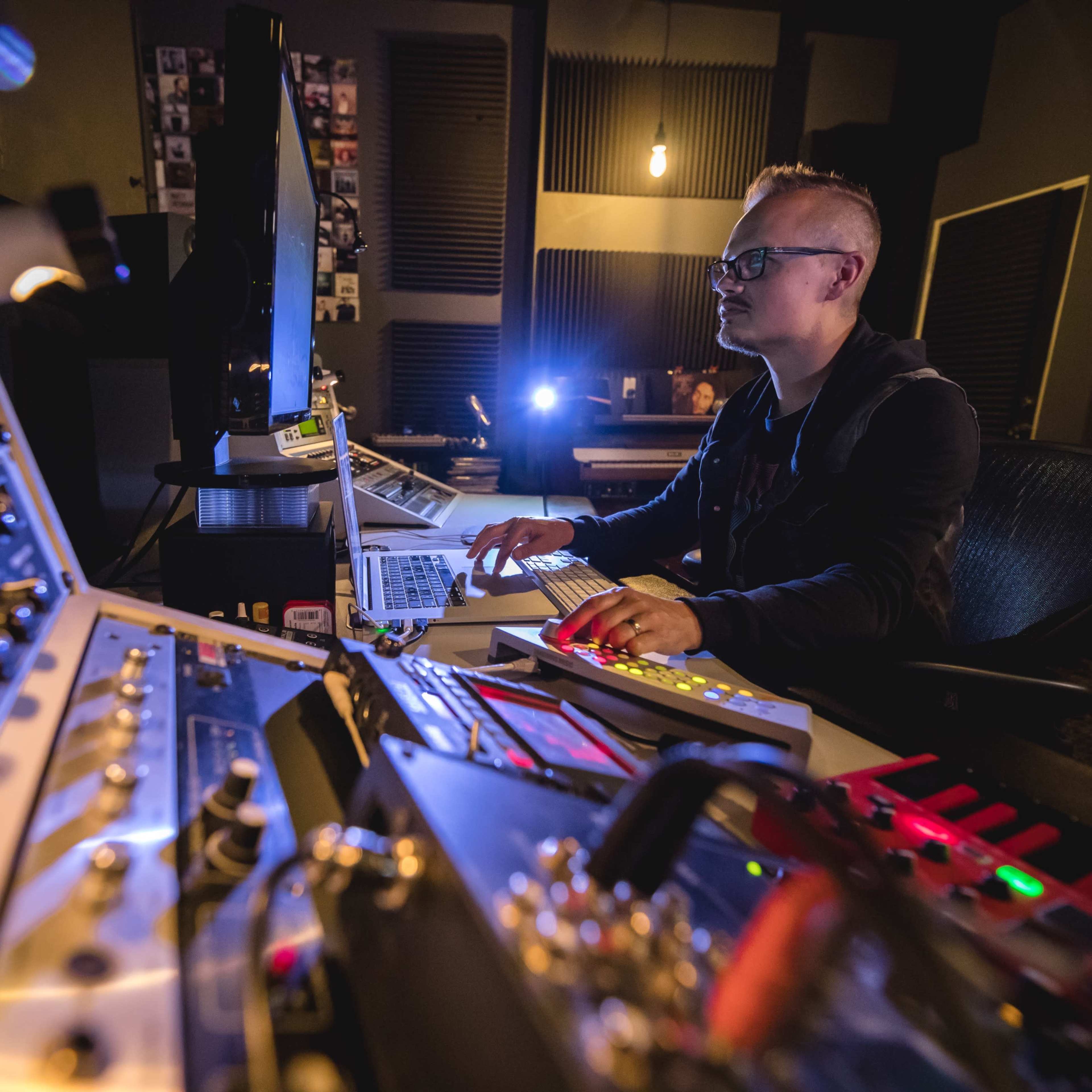 A person works at a music production console surrounded by audio equipment and a computer in a recording studio.
