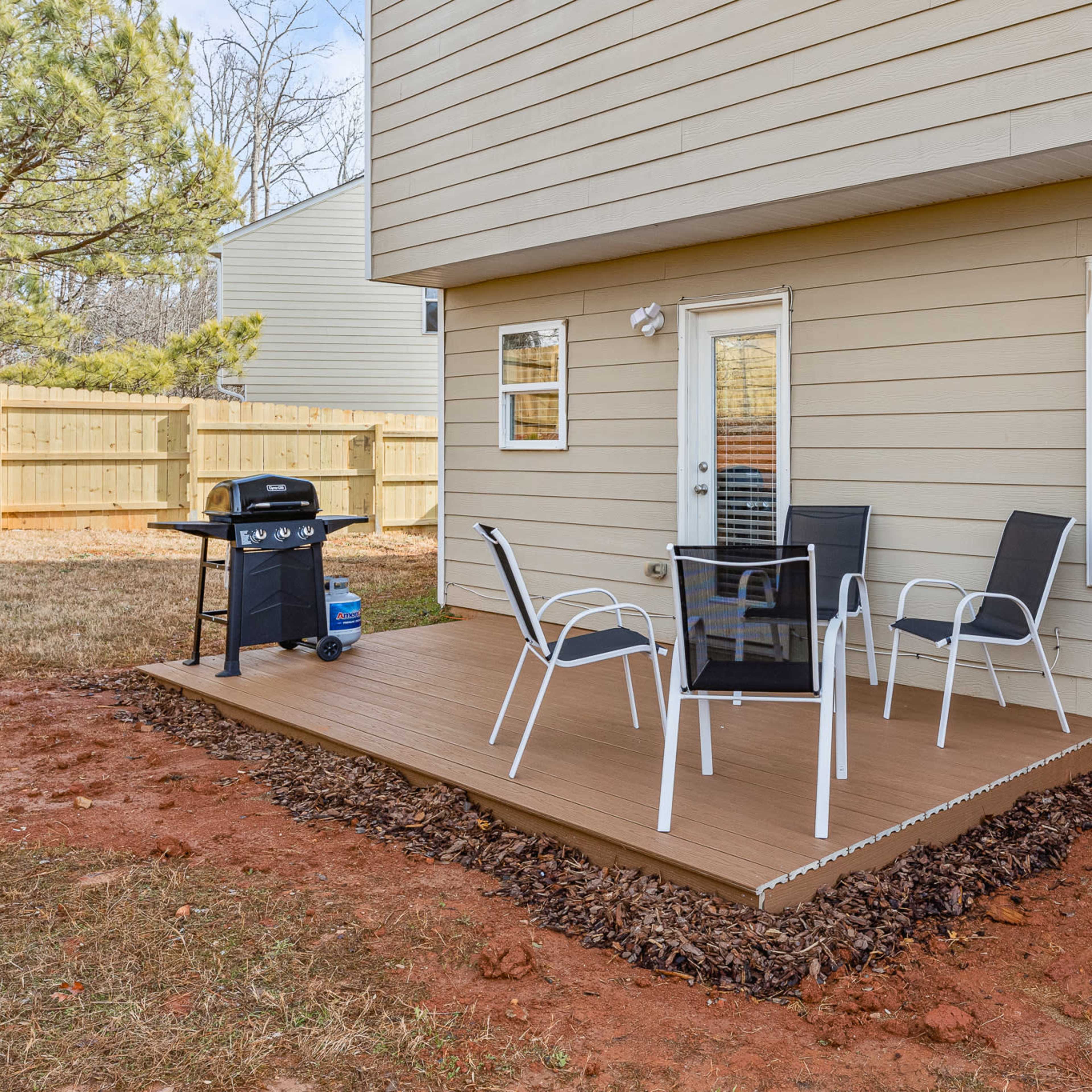 A small deck with a table and chairs, along with a charcoal grill, adjacent to a beige house in a grassy yard.