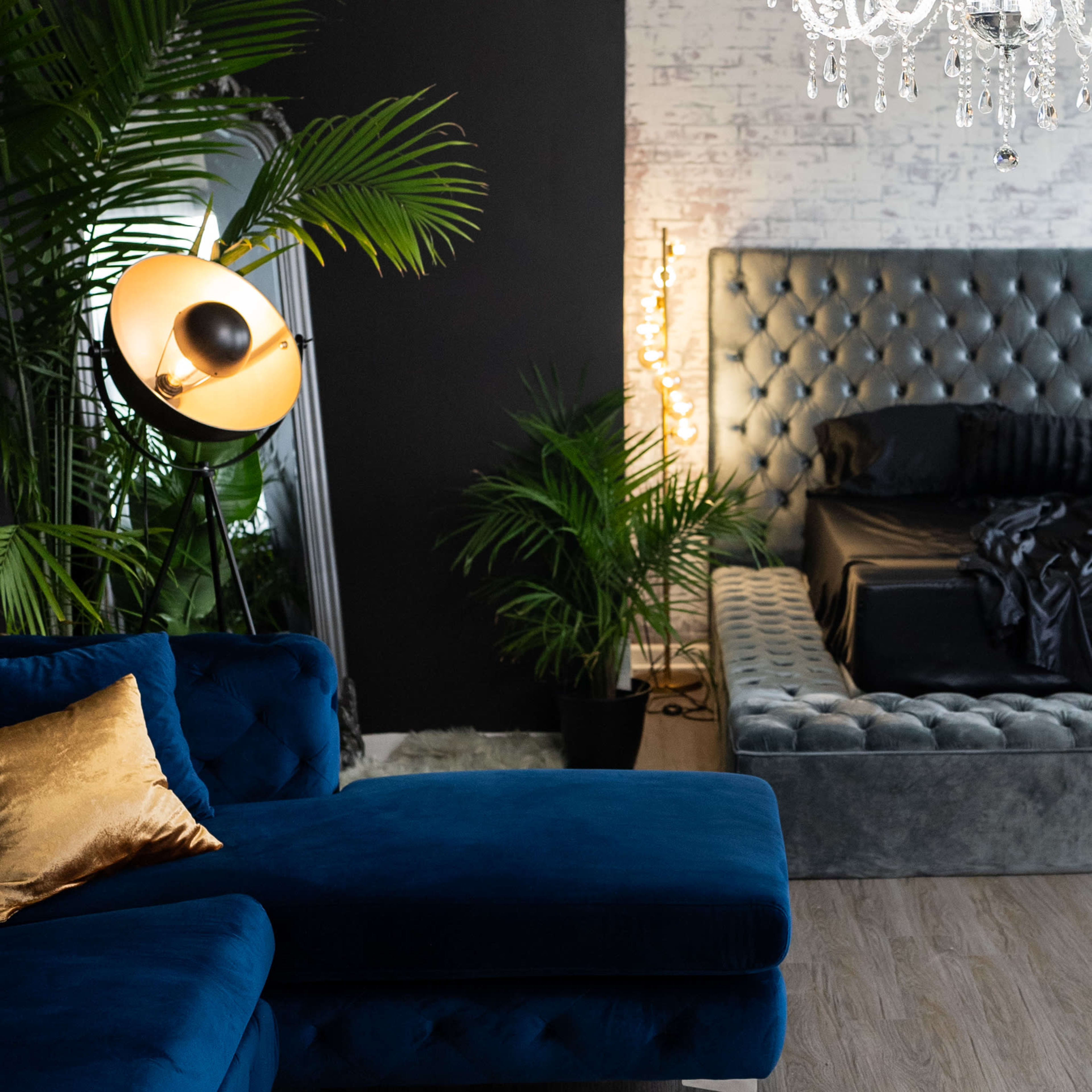 A modern living room features a blue sectional sofa, a crystal chandelier, and a tufted bed framed by tall plants.