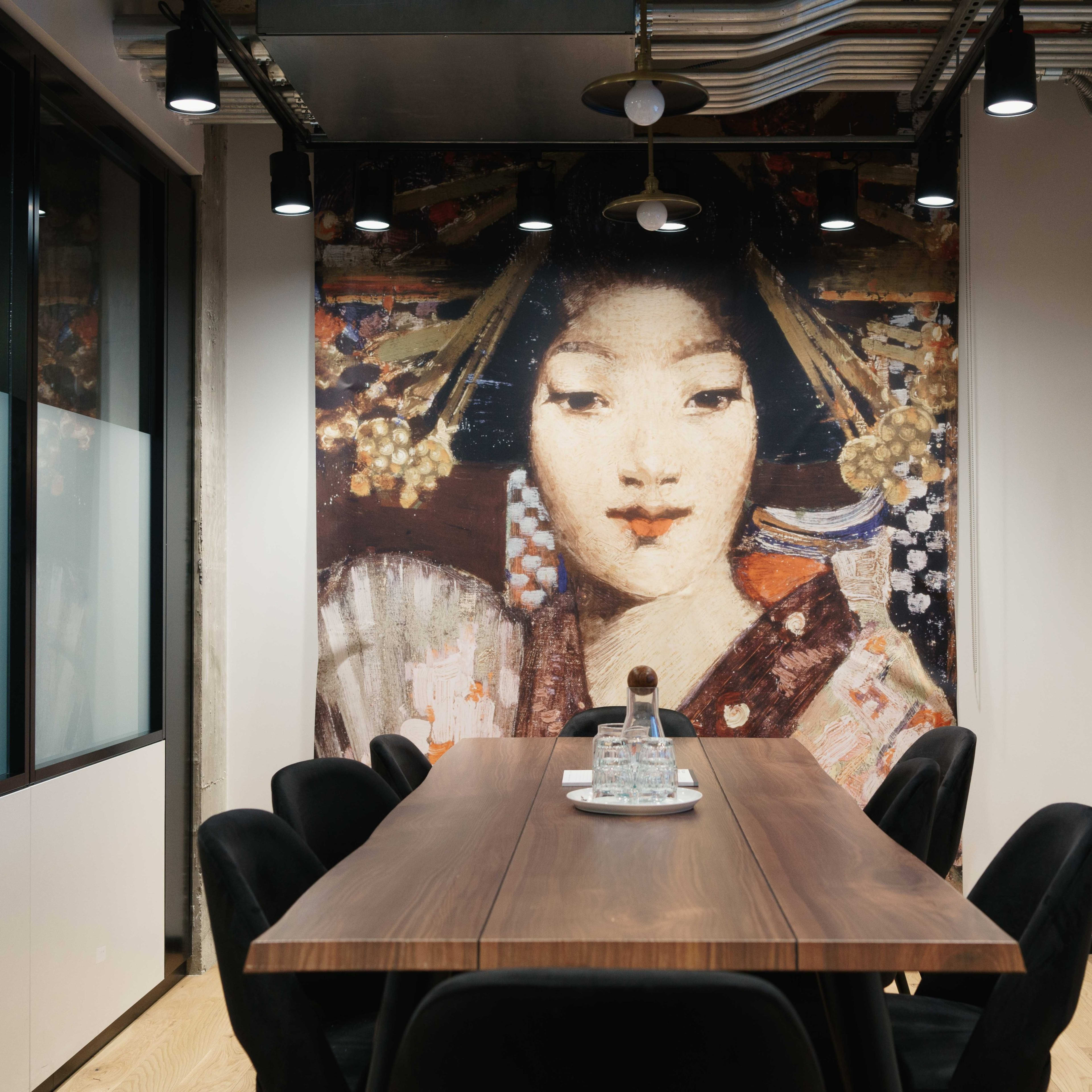 A conference room features a long wooden table surrounded by black chairs, with a large mural of a woman in traditional attire on the wall behind.