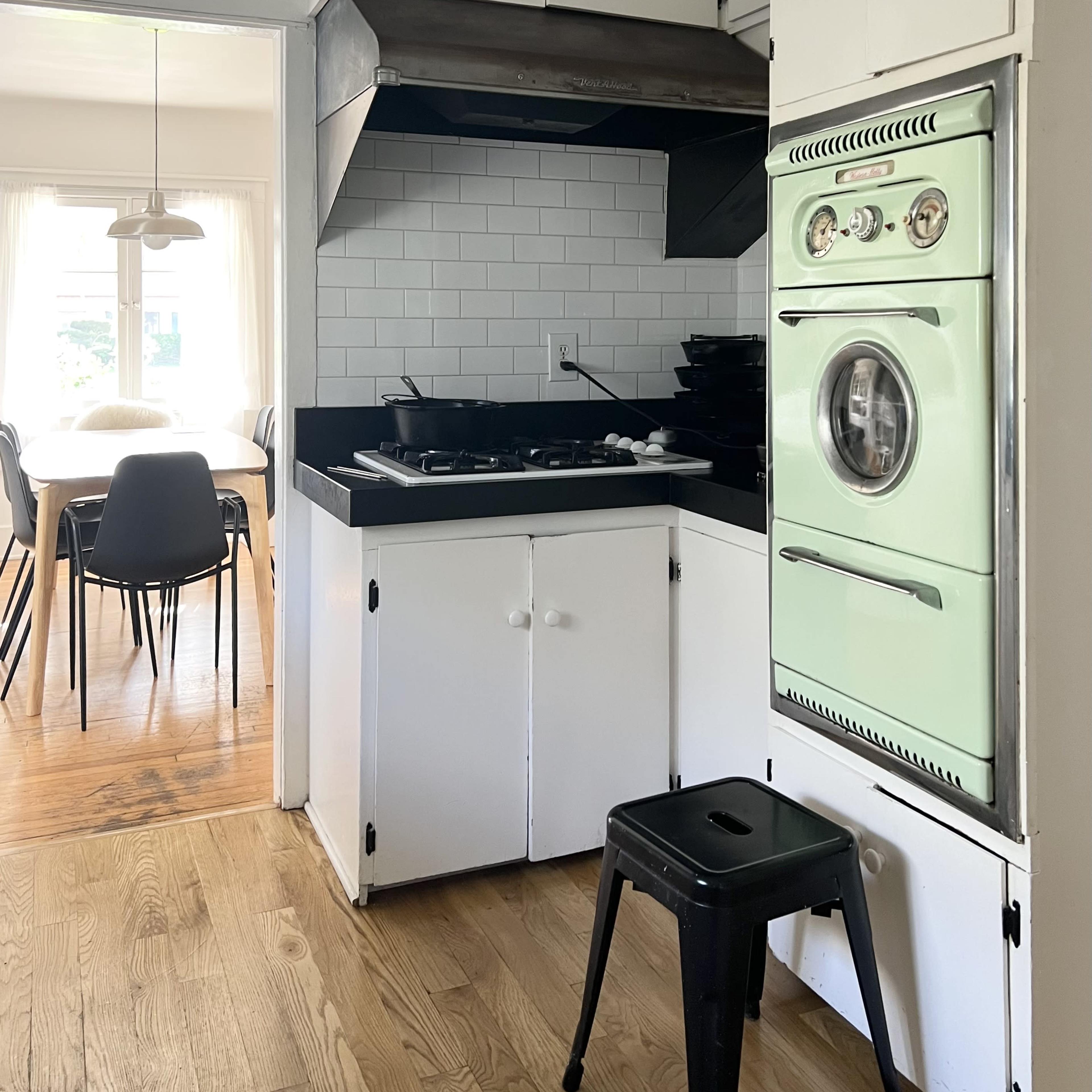 A compact kitchen with white cabinetry, a stainless steel range hood, and a mint-green retro oven beside a black countertop, leading to a dining area in the background.
