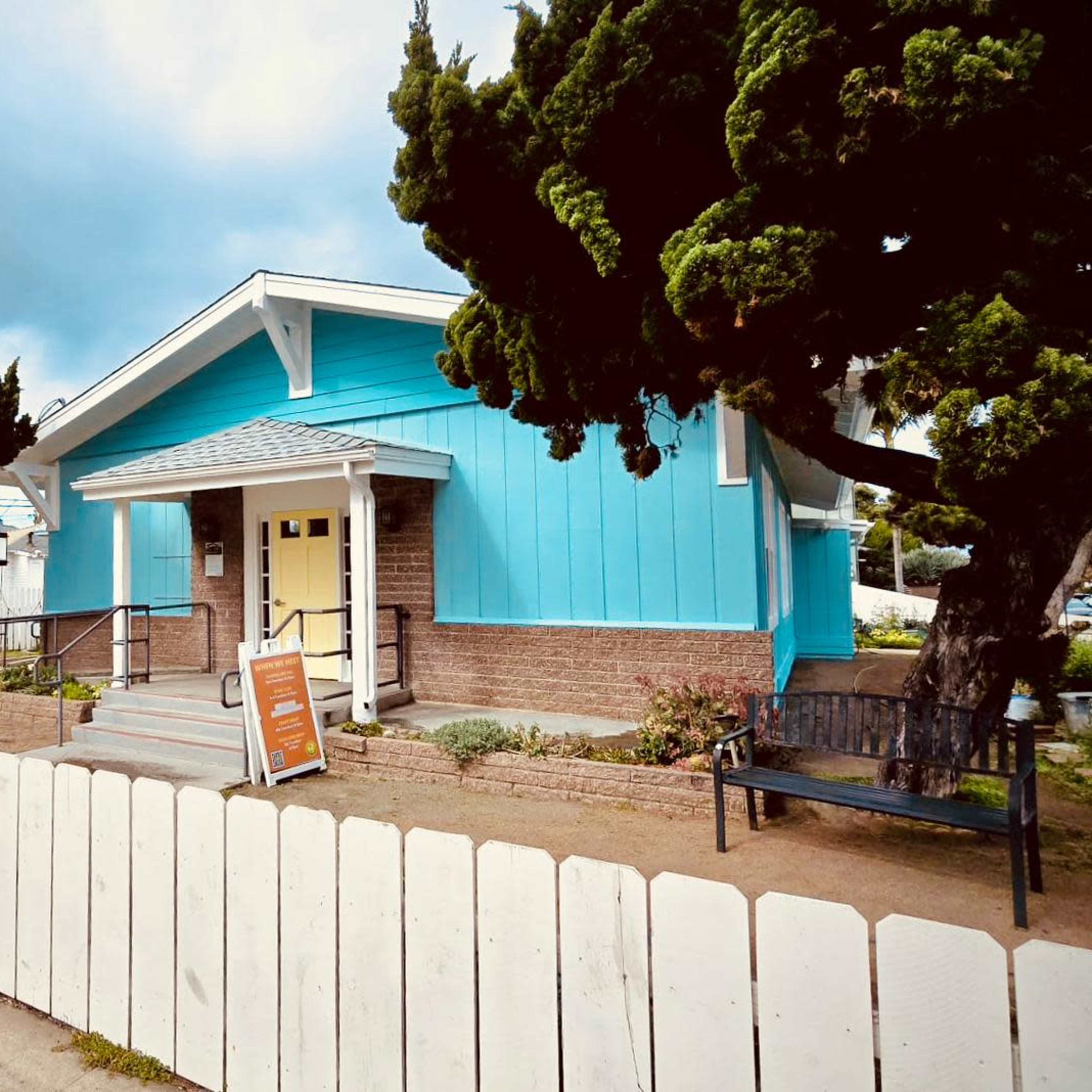 A blue house with a front porch and a white picket fence stands among trees and shrubs.