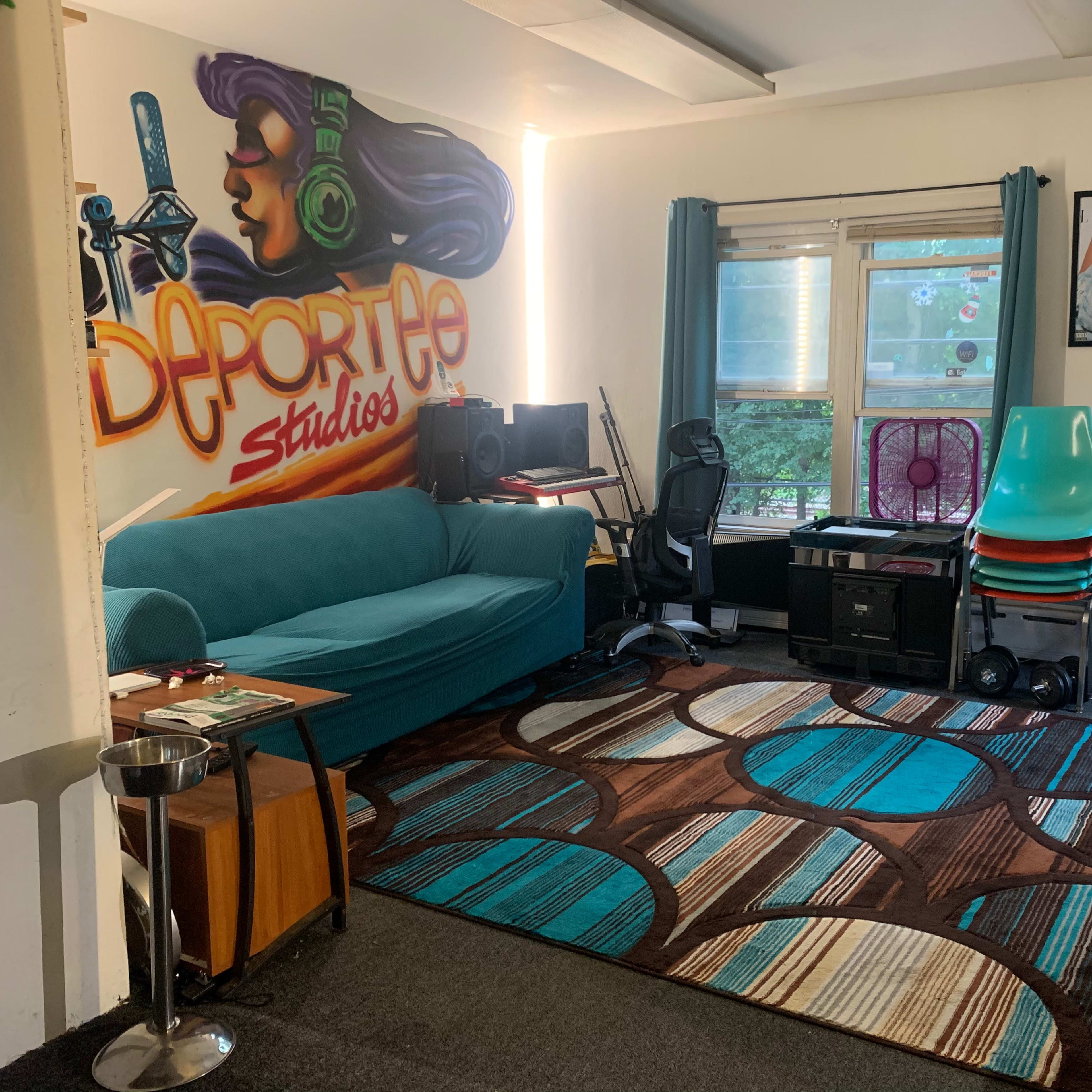 A room featuring a teal couch, a large area rug with circular designs, a mural with the text "Deportee Studios," and various furniture items including stacked chairs and a small table.