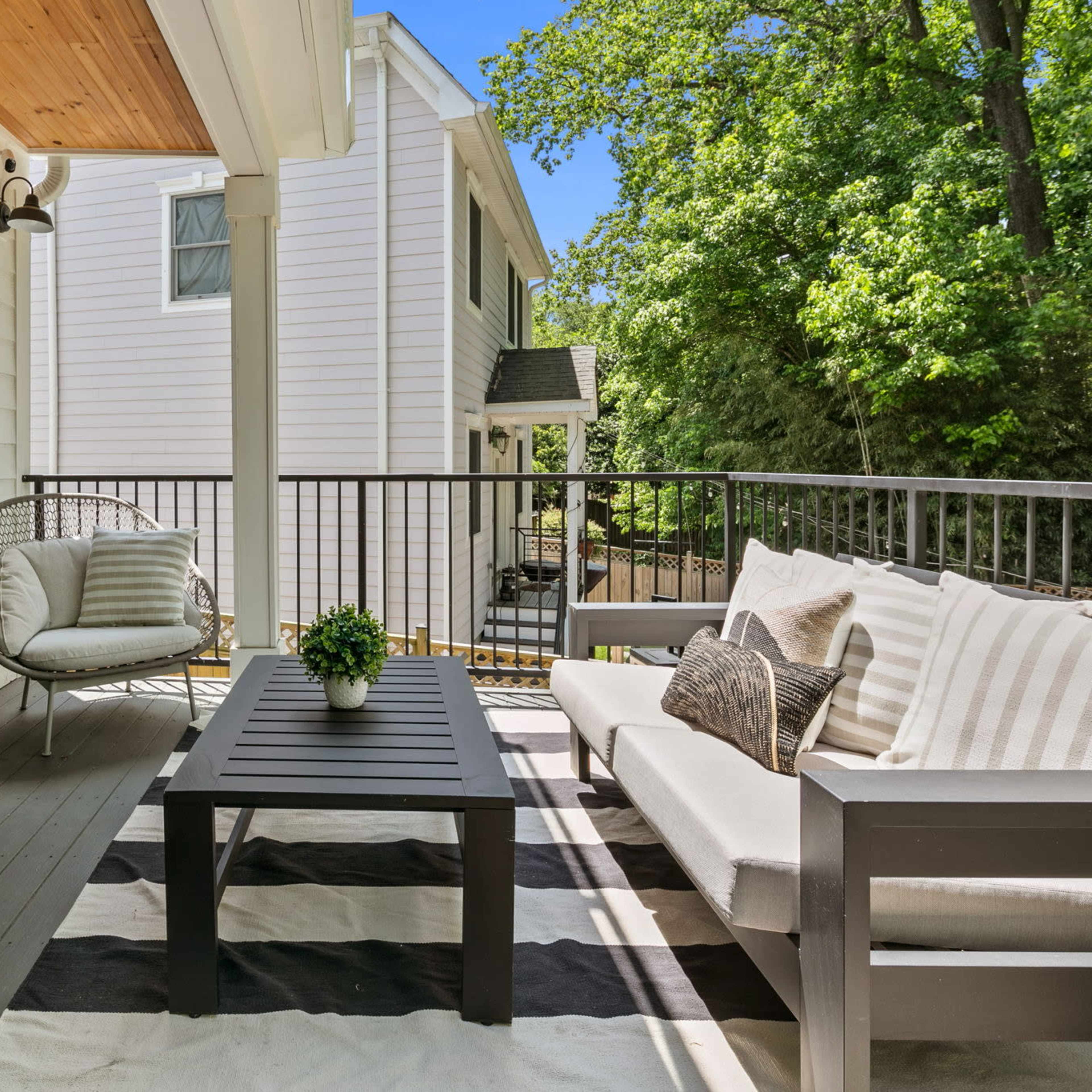 A modern balcony features a seating area with a striped rug, a coffee table, and greenery surrounded by trees.