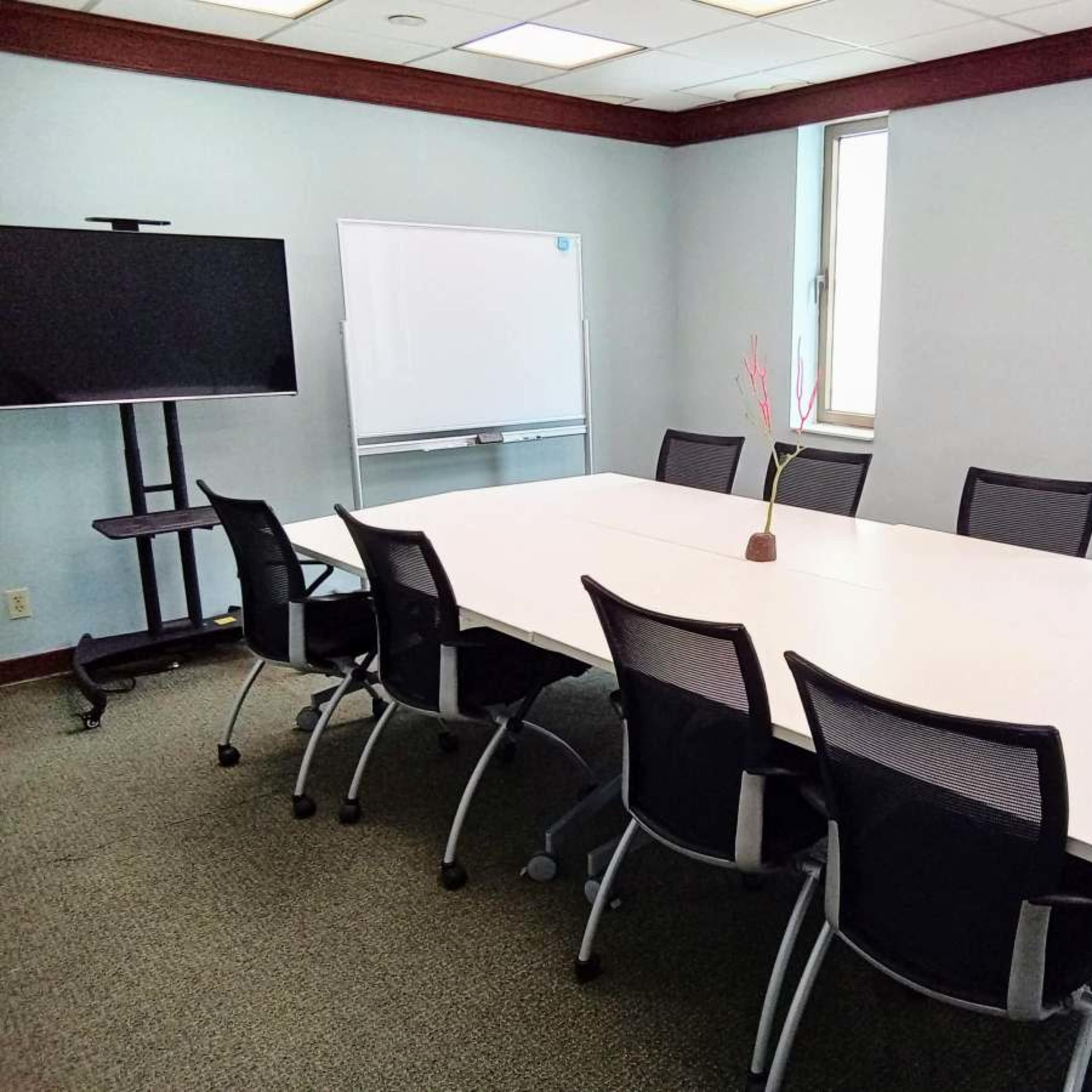 A conference room features a large white table surrounded by eight black chairs, with a television on a stand and a whiteboard in the background.
