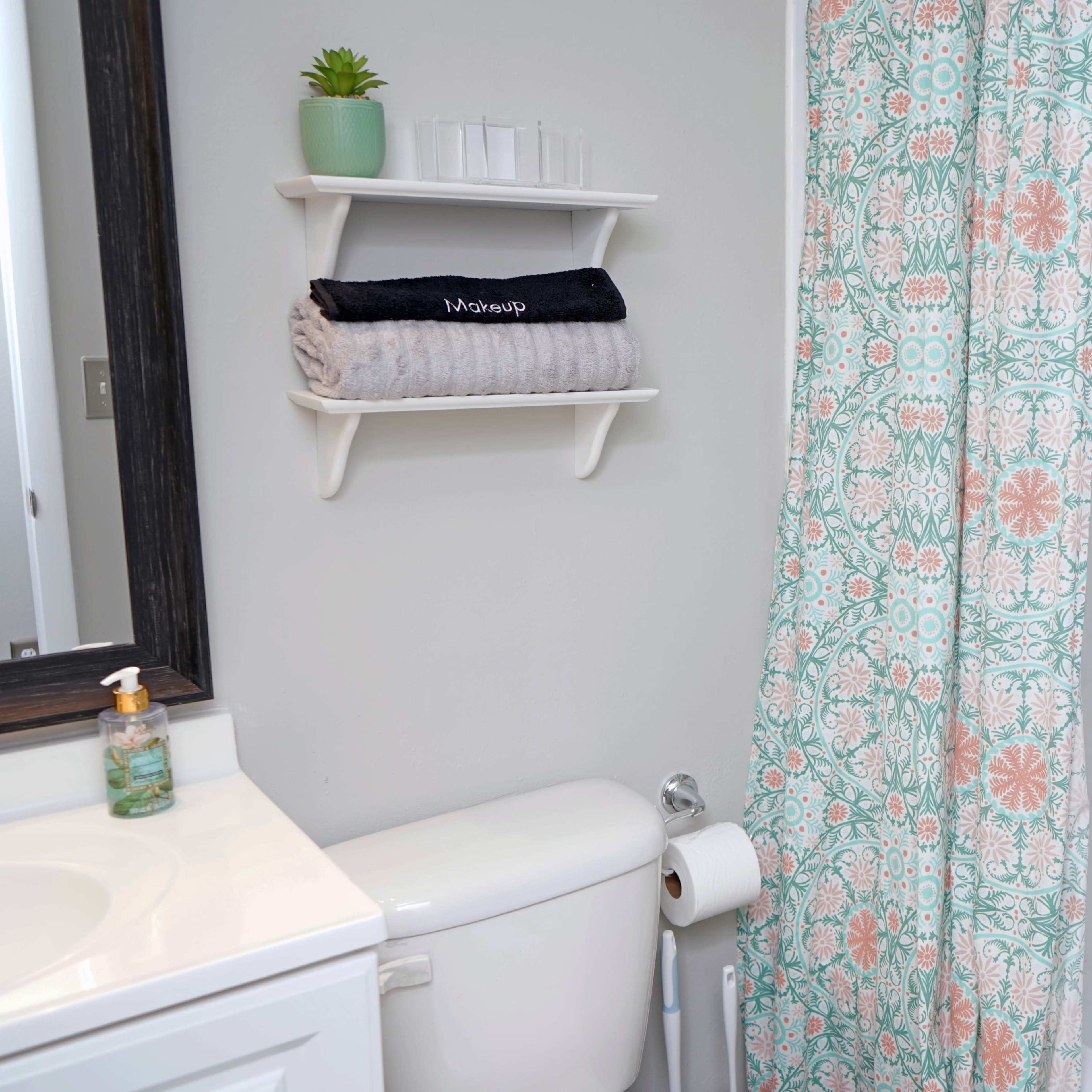 A bathroom with a shower curtain featuring a floral pattern, a shelf with towels, and various toiletry bottles on the bathtub ledge.