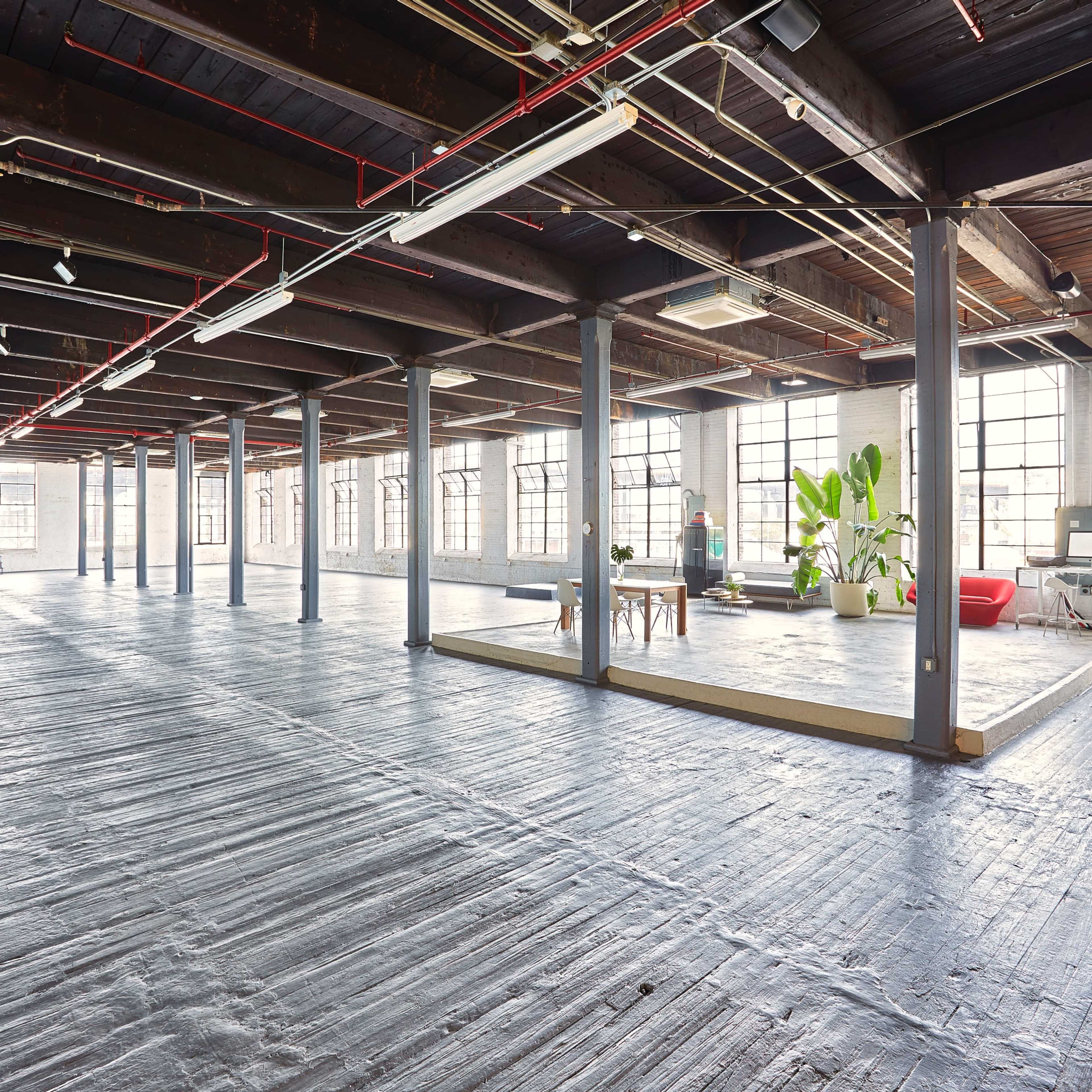 A spacious, open-concept loft features large windows, wooden beams, and polished concrete flooring, with distinct areas for lounging and working.