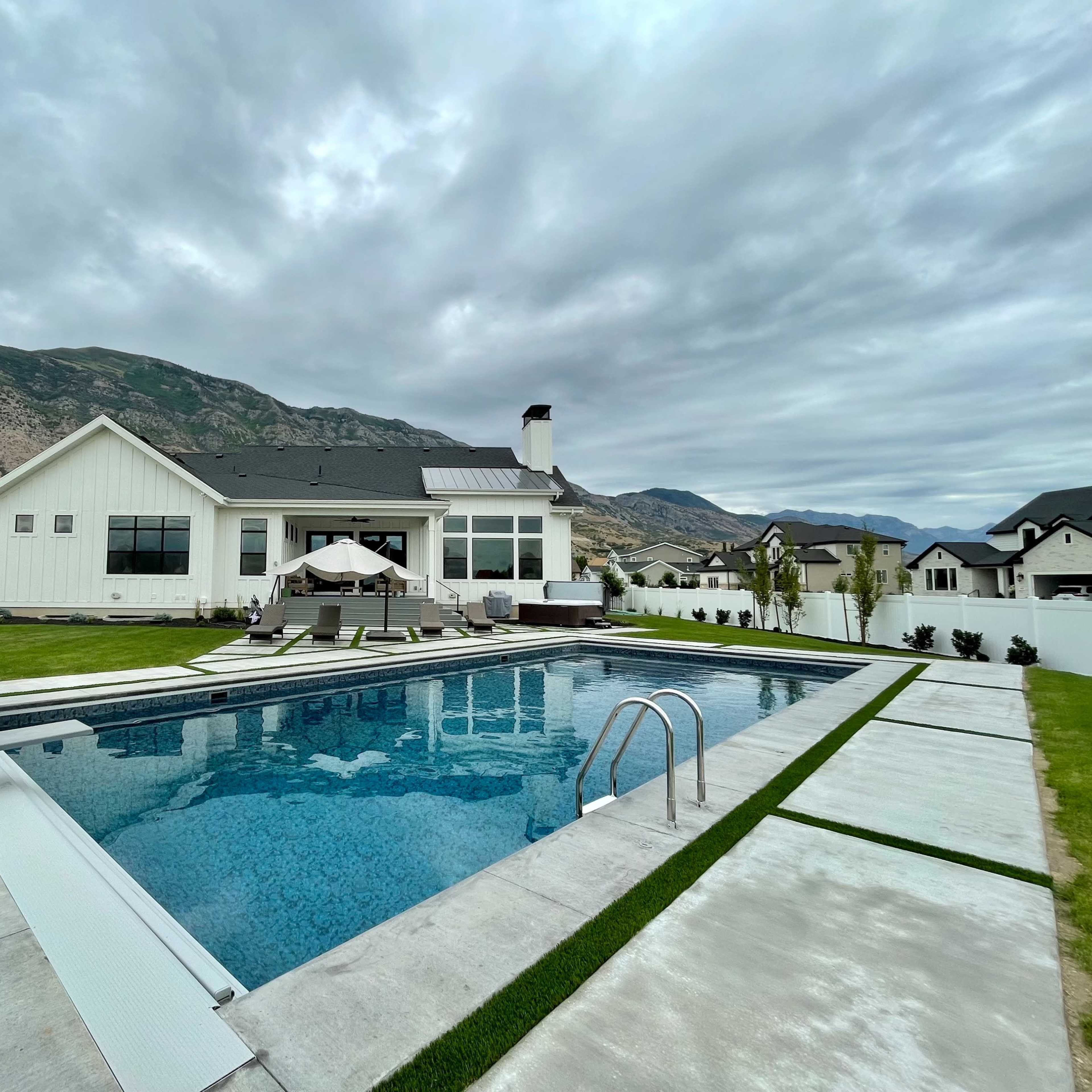 A modern house with a landscaped backyard featuring a swimming pool, lounge chairs, and a mountain backdrop under overcast skies.
