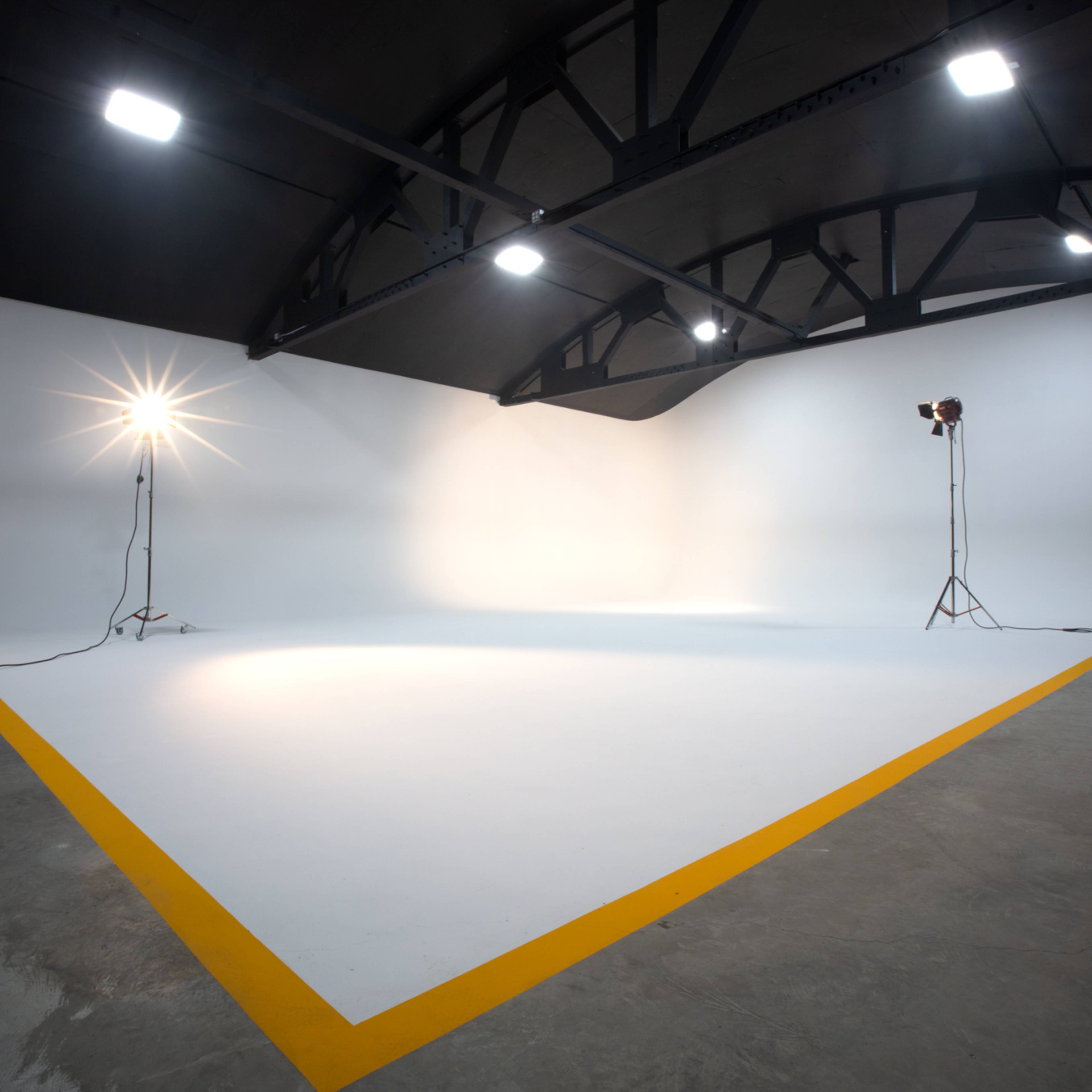 A spacious photography studio with a white backdrop, yellow corner markings, and two lighting stands positioned on either side.