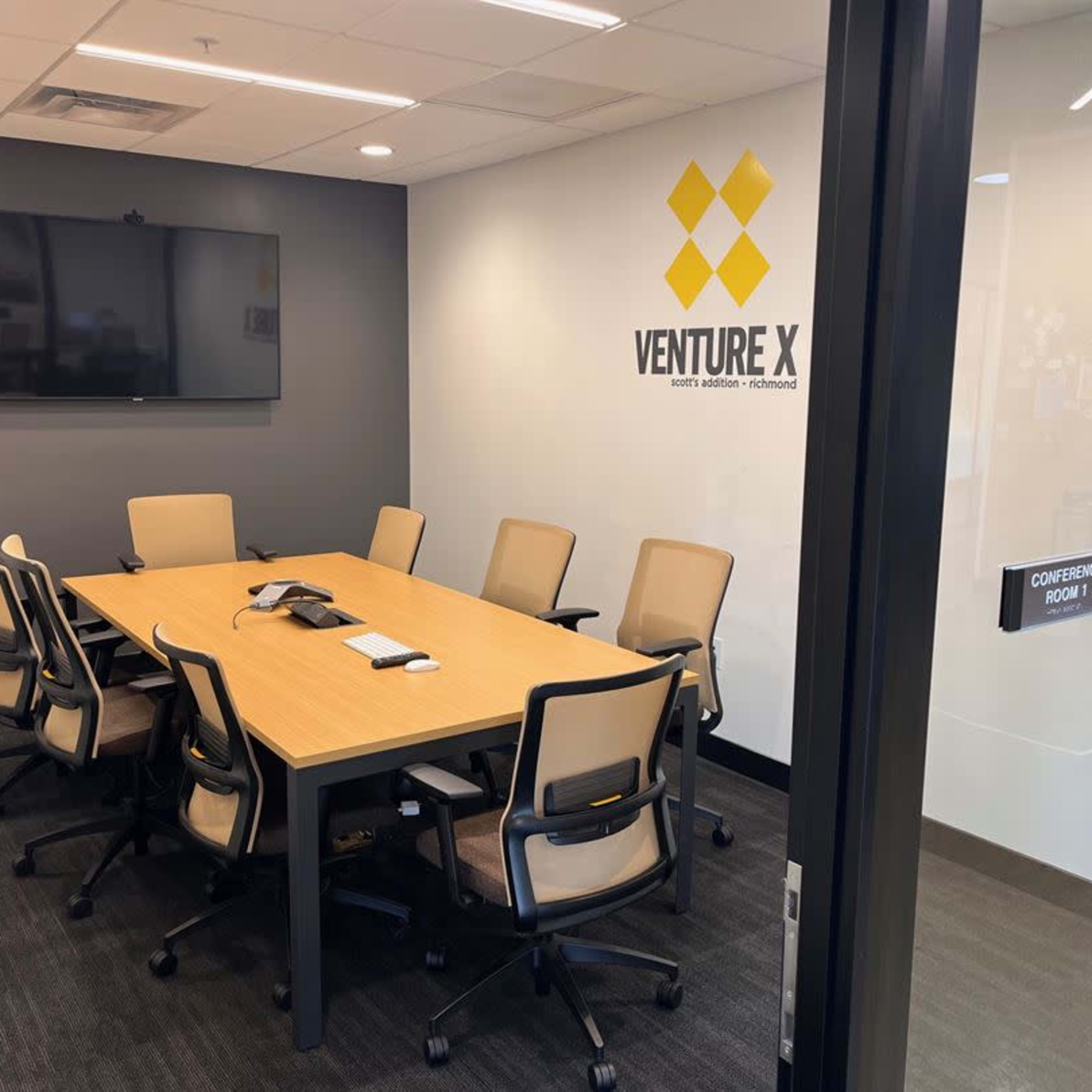 A conference room equipped with a long table, eight chairs, a large wall-mounted screen, and a phone on the table.