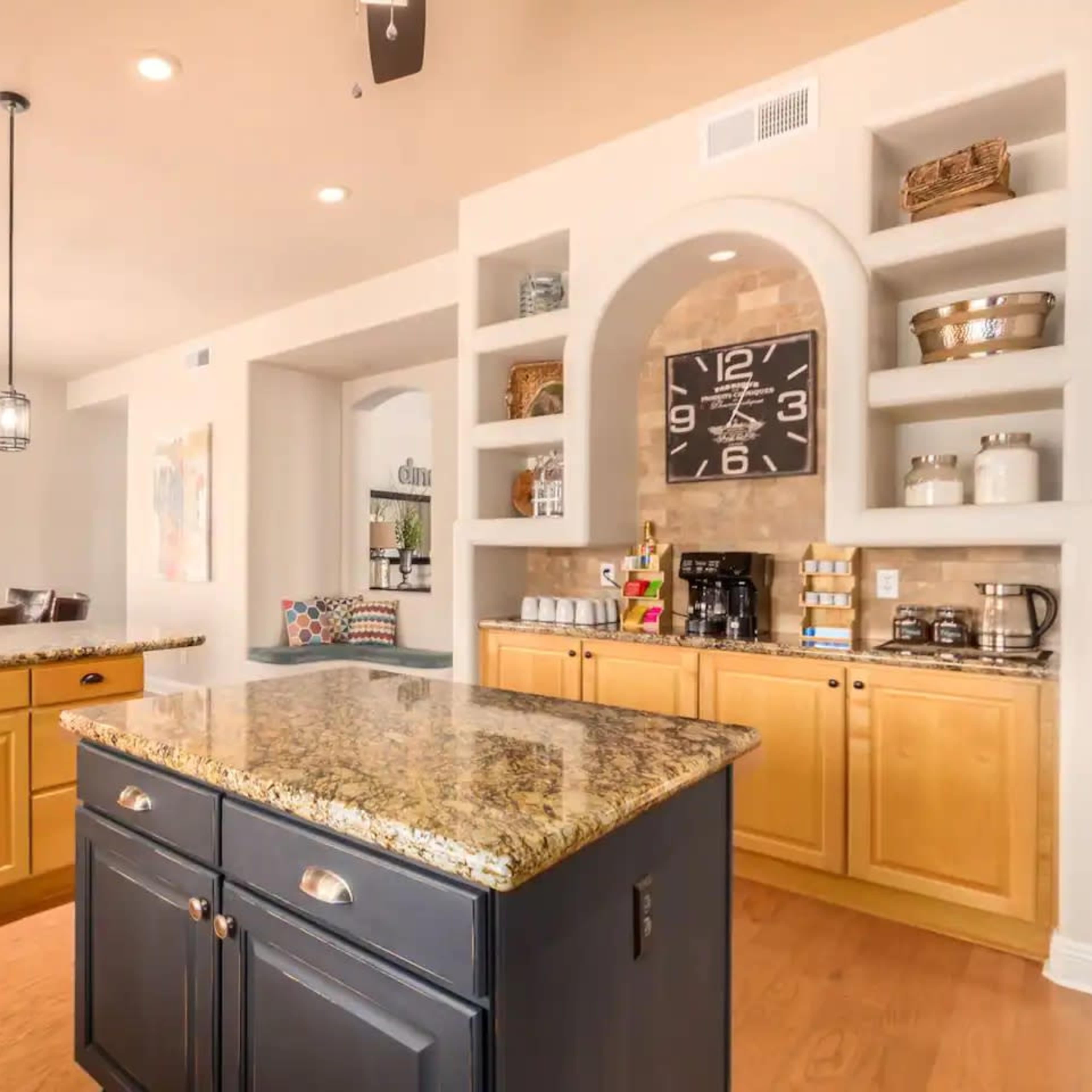 A modern kitchen with a central island featuring a dark countertop and light cabinetry, set against a wall with decorative shelves and a large clock.