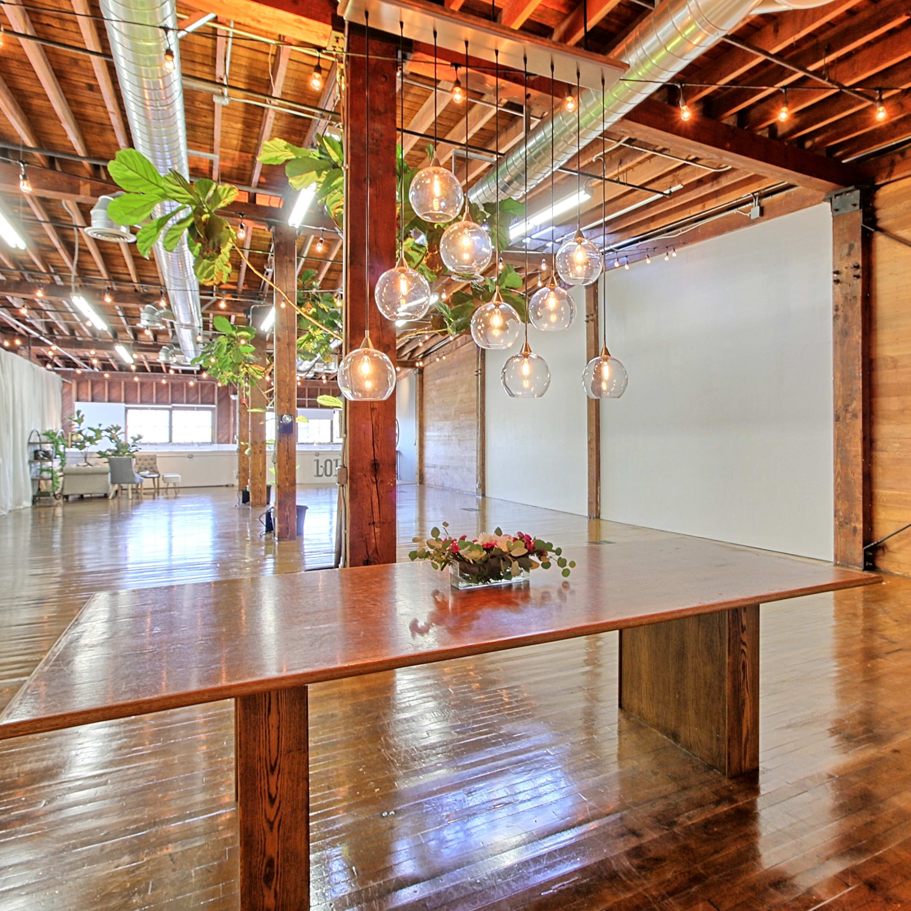 A spacious room with wooden beams, polished floors, and a large table adorned with a floral centerpiece under hanging glass light fixtures.