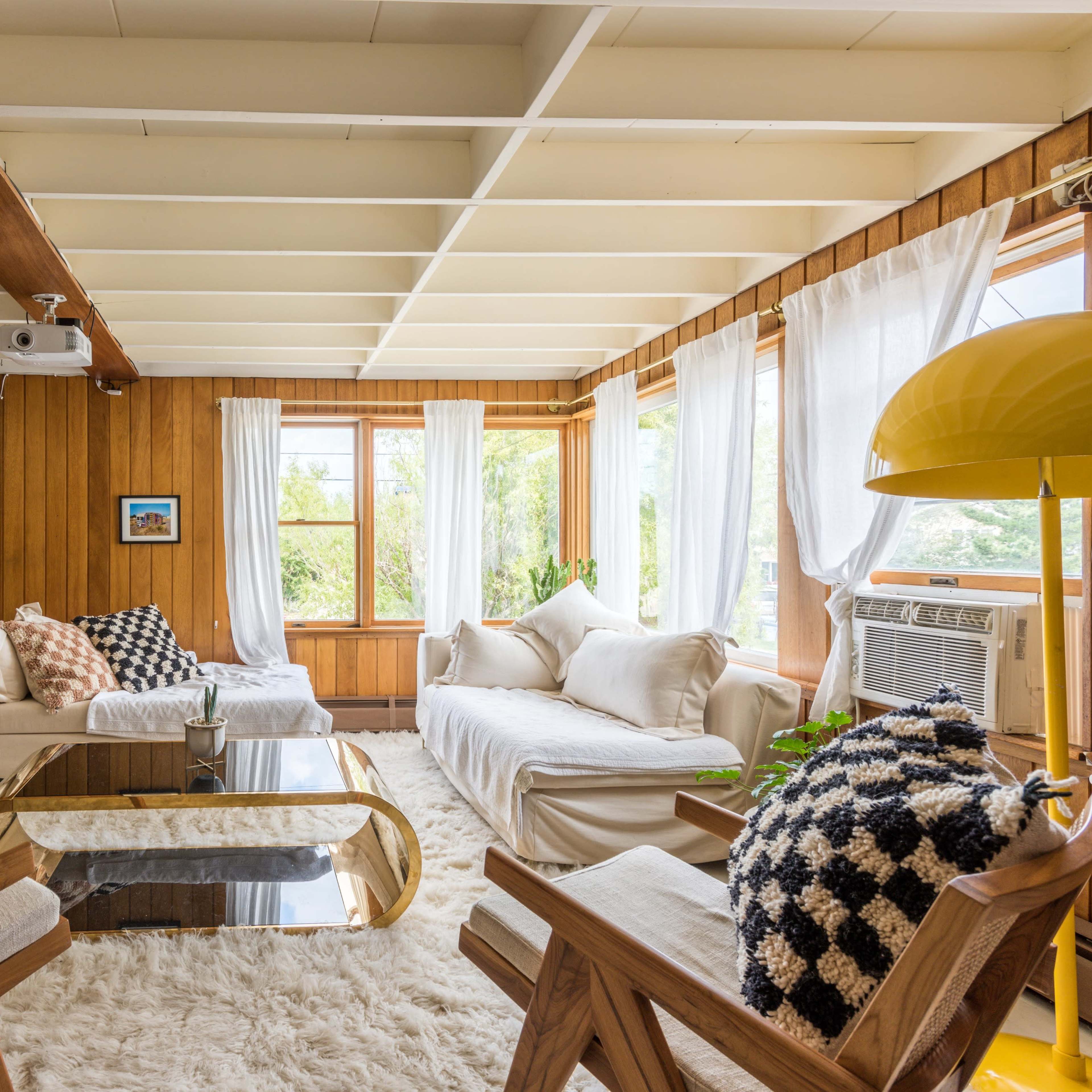 A bright living room features a white sofa, wooden chairs, a glass coffee table, and large windows with sheer curtains, surrounded by potted plants.