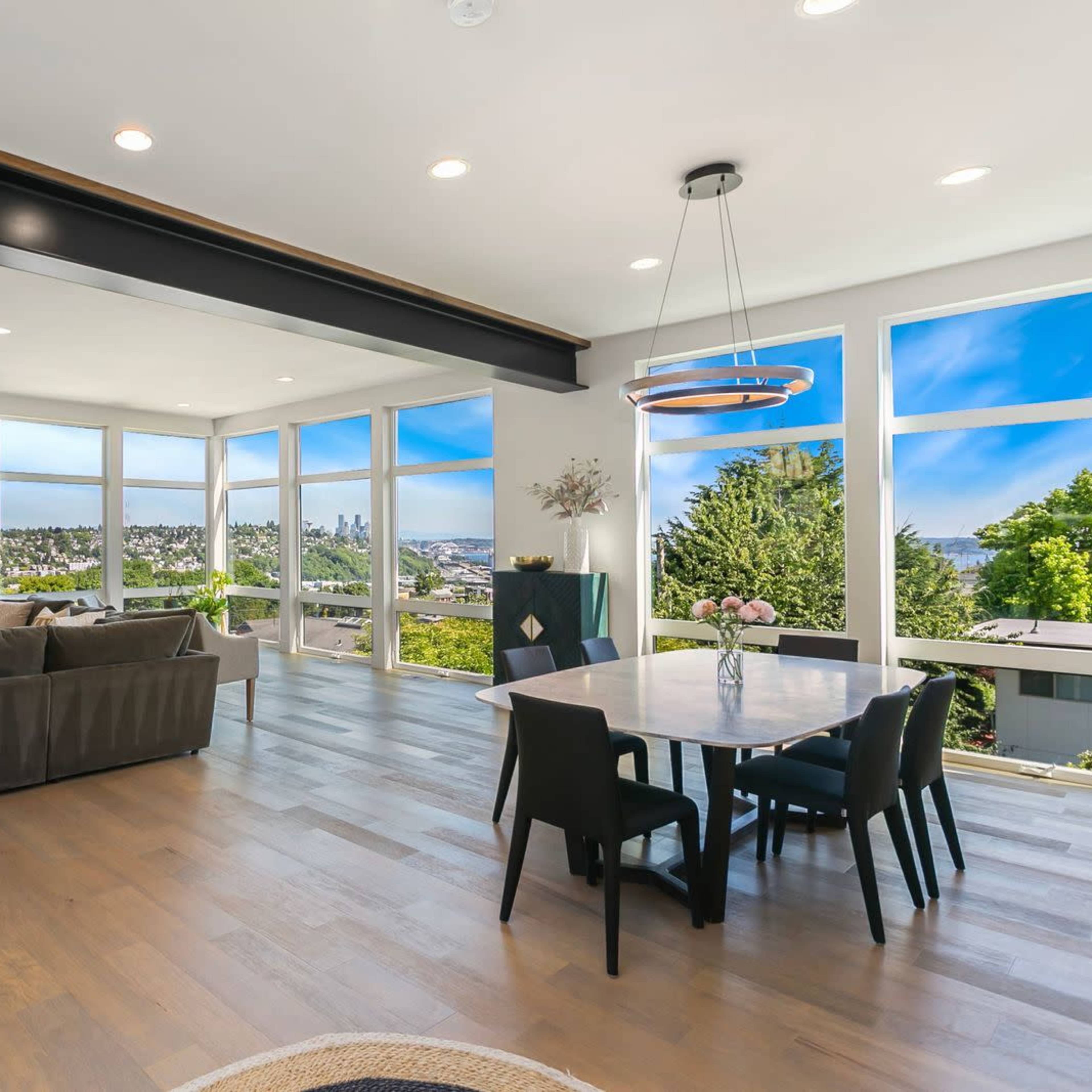A modern living space with large windows, a gray sofa, a dining table set for six, and a view of the landscape outside.