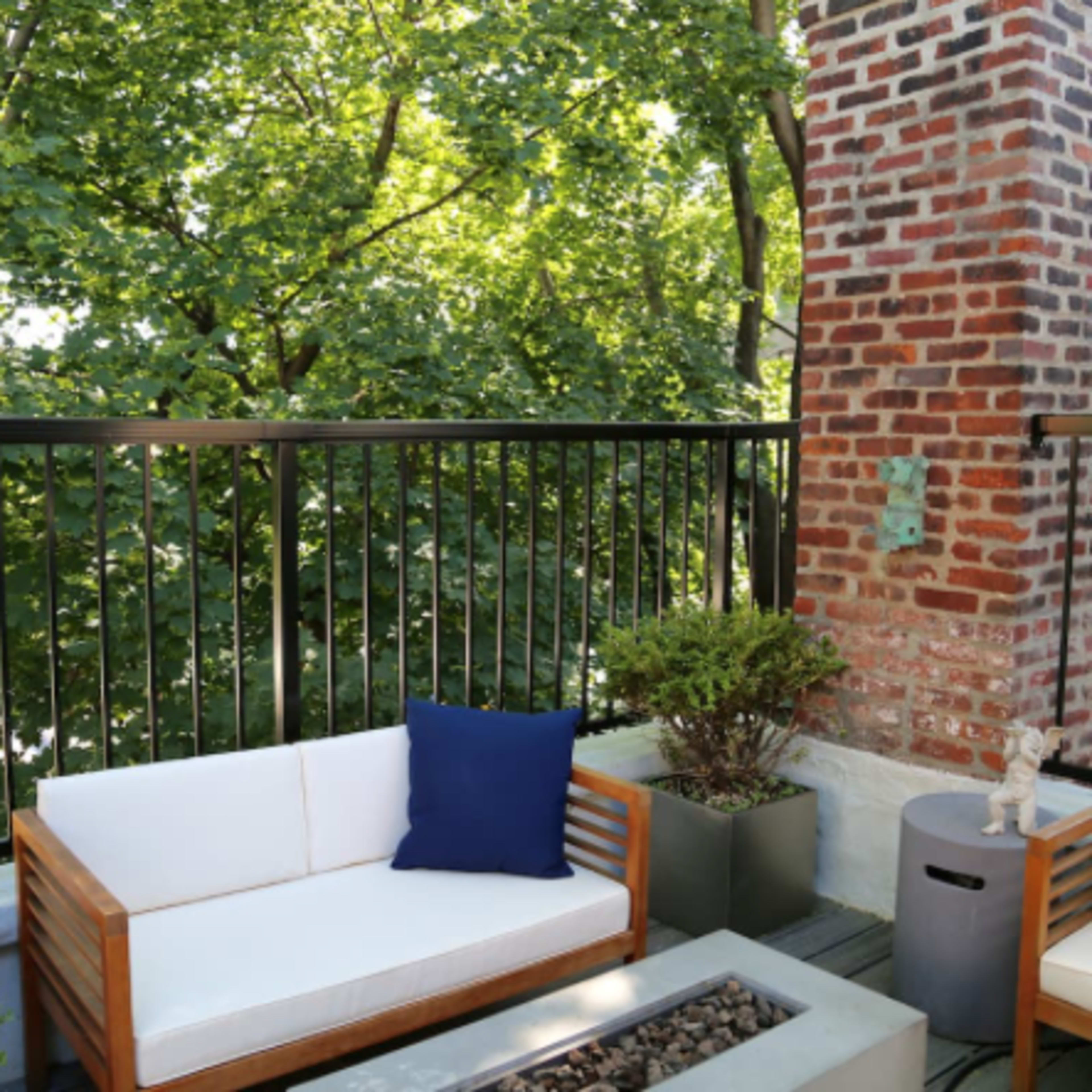 A balcony features two wooden chairs with white cushions and blue pillows, alongside a small table with a stone fire pit, surrounded by lush green trees.