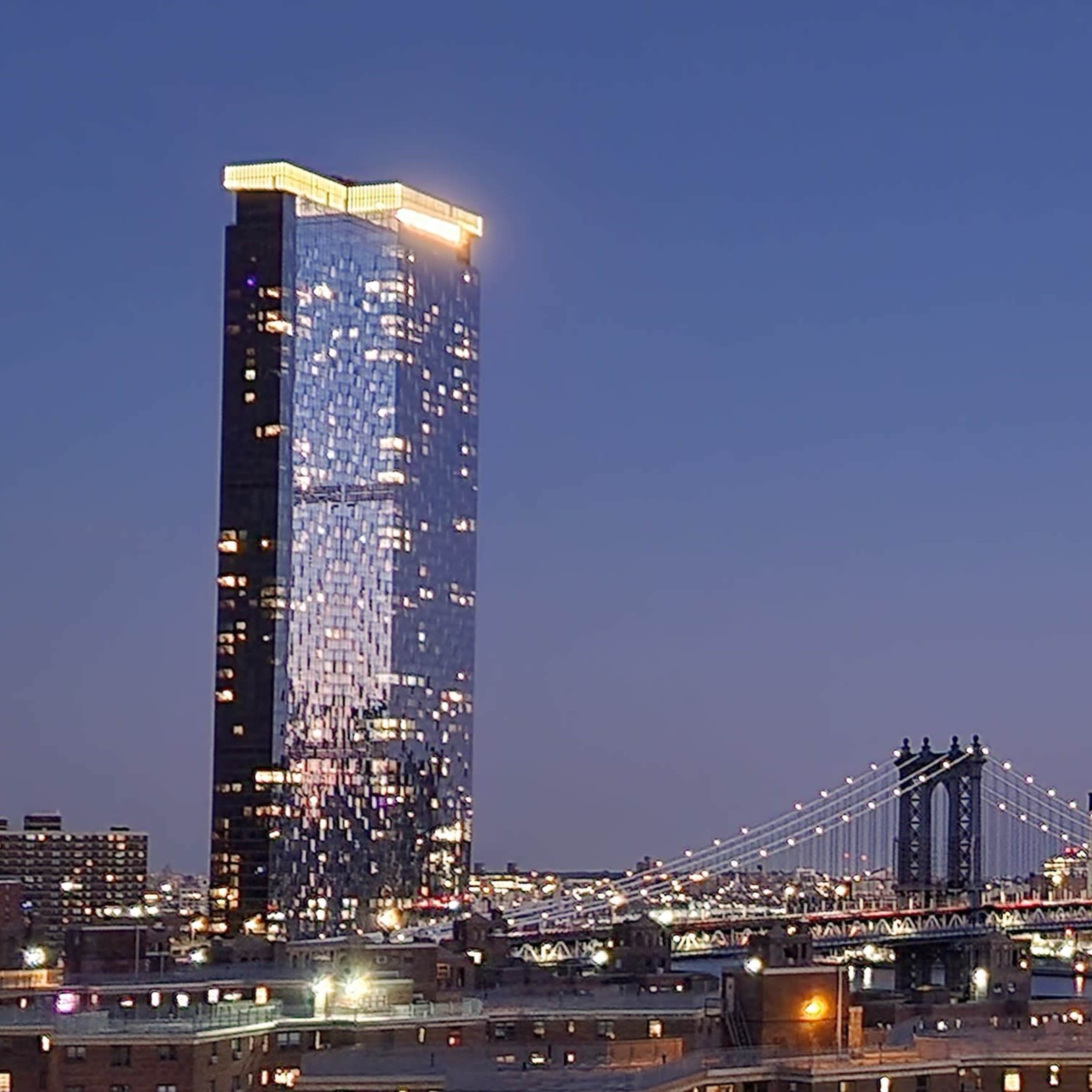 A skyline view of illuminated skyscrapers and the Brooklyn Bridge at dusk, with a clear sky transitioning from daylight to night.